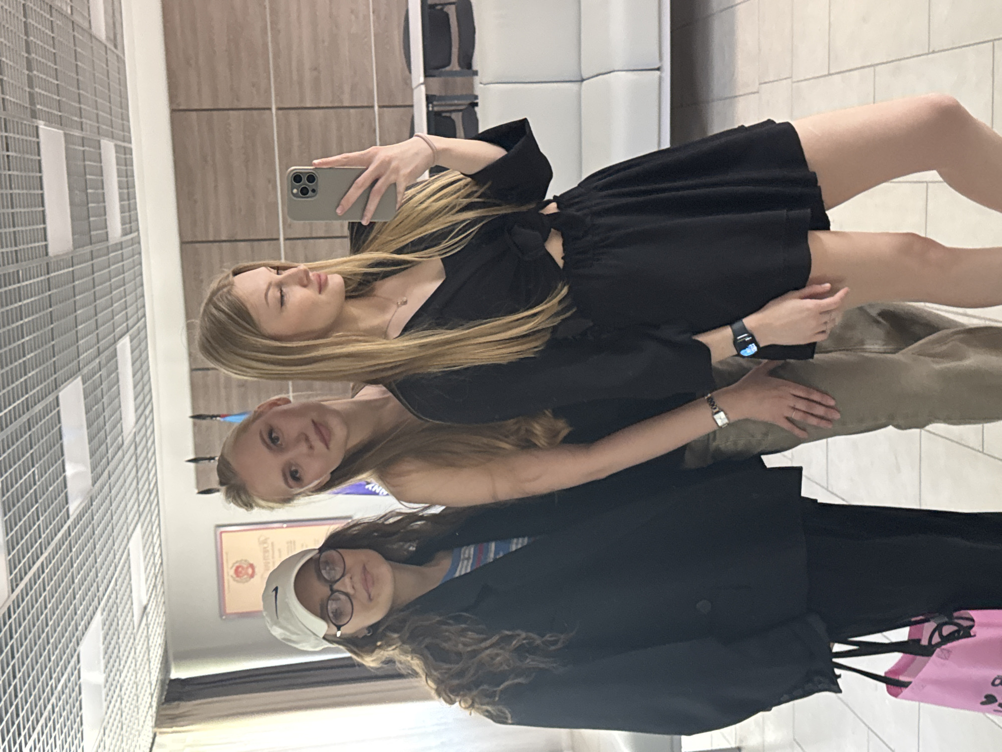 Three young women are posing for a selfie in what appears to be an interior space. The woman in the forefront wears glasses and a light-colored cap, dressed in a black blazer, with curly hair. The woman in the middle has light brown hair and is wearing a neutral-toned top. The third woman, at the top of the image, holds a smartphone to take the picture. She has long blonde hair, is wearing a black dress, and has a wristwatch on her left wrist. The background includes a wall with horizontal panels and what looks like a tiled wall. A white upholstered seating area is visible in the upper part of the image. Based on available visual information, it's not possible to determine the exact location in Perm, Russia.