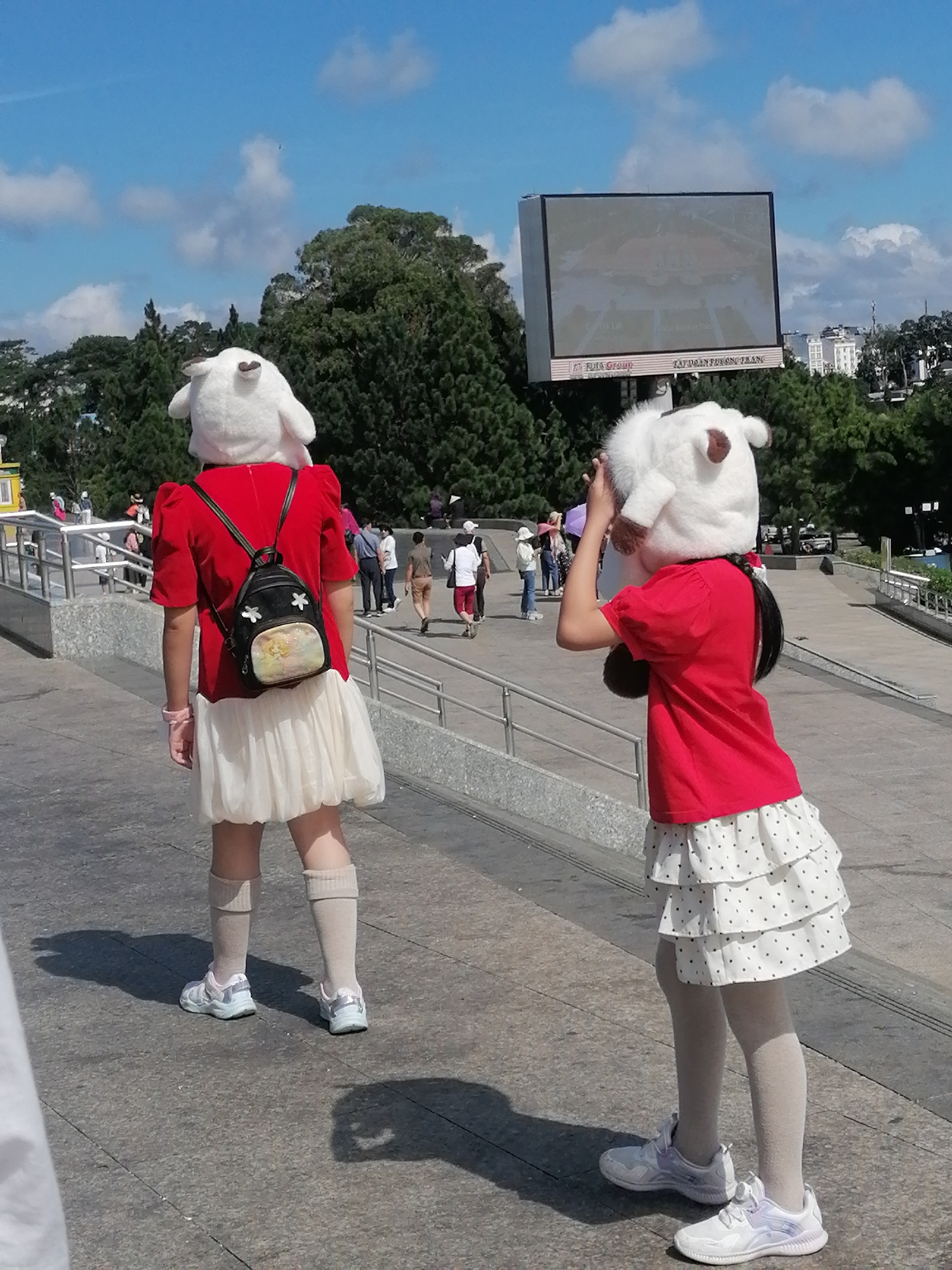 The image depicts two individuals, likely children, dressed in red tops and white, layered skirts. Both are wearing large, white plush animal head costumes, resembling sheep or lambs, with brown ear accents. The individual on the left also wears a small black backpack adorned with floral designs and a cartoon character. The individual on the right holds one of the costume's ears near their face. They are standing on a paved pathway, which appears to be part of a public space with tiered seating or platforms. In the background, trees, buildings, and a large outdoor LED screen displaying an image can be seen. The setting is outdoors, under a partly cloudy blue sky, and is consistent with a location in Da Lat, Vietnam, a city known for its hilly terrain and lush greenery. The overall scene suggests a casual outing or event.