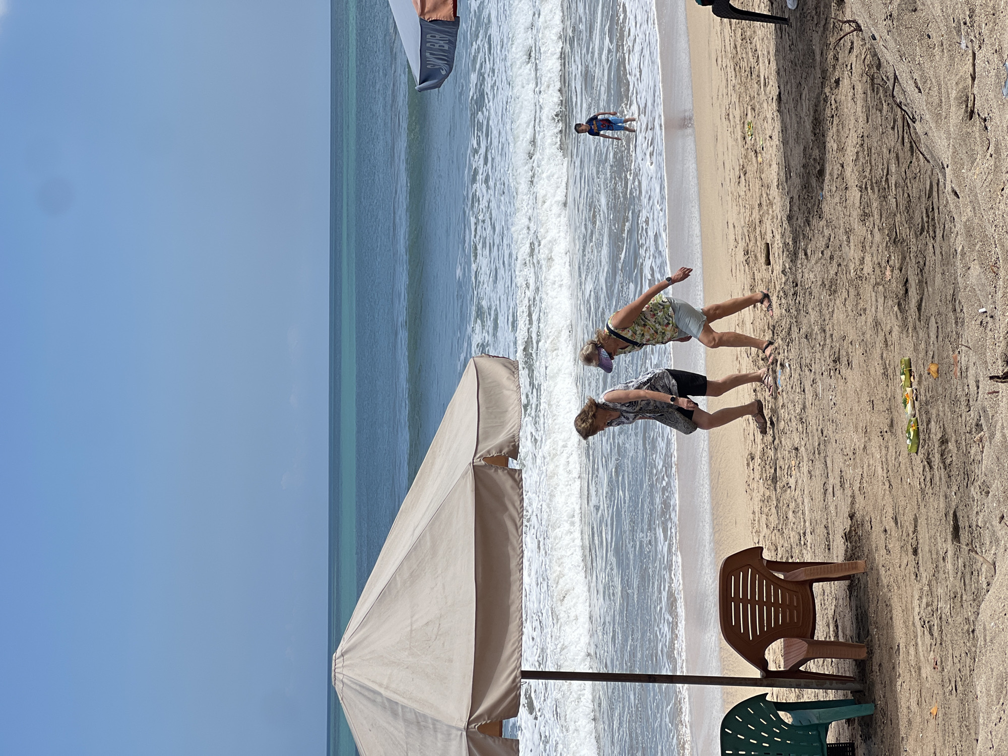 In Kuta, Indonesia, two women walk on a sandy beach. The woman on the left wears a black and white patterned sleeveless top and black shorts. The woman on the right wears a floral print sleeveless top and light-colored shorts. A young boy in a dark t-shirt and blue shorts stands in the shallow ocean water. A large, tan umbrella stands on the left side of the beach, casting a shadow. In the background, the turquoise ocean meets the clear blue sky. Several plastic chairs are scattered on the sand, including a green one on the left and a brown one in the center. Small offerings, possibly Balinese canang sari, are visible on the sand near the right of the frame. The overall scene depicts a typical beach day with people enjoying the water and the sun.