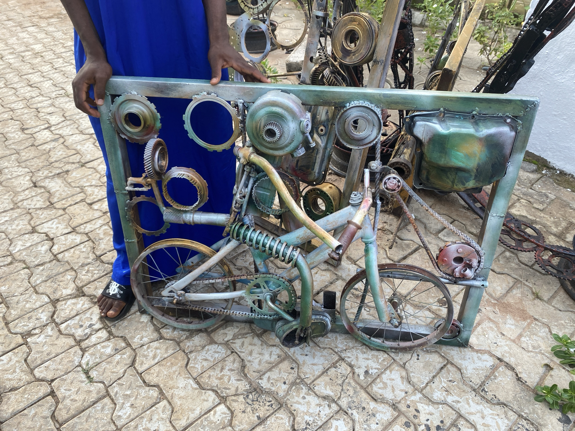 In an outdoor setting with a paved ground made of light-colored interlocking stones, a person wearing a bright blue garment and black sandals is holding a large, rectangular metal sculpture. The sculpture is a creative assemblage of recycled metal parts, primarily gears and bicycle components, forming the shape of a bicycle within its frame. The bicycle appears to be made of various salvaged materials, painted in shades of green, blue, and rust. The frame of the sculpture itself is also constructed from metal, painted in a similar color palette. The background shows a white wall and some greenery. The person's hands are visible, one resting on the top edge of the sculpture and the other near the gears of the bicycle. The sculpture is the main subject, with the person acting as a presenter or owner.