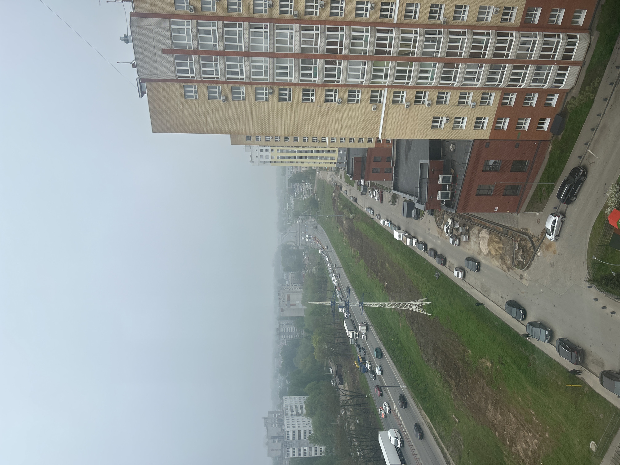 The image shows a high-angle view of an urban landscape on a foggy day. Prominent features include a tall, yellow and red brick apartment building with numerous windows, a road with cars parked and moving along it, and a green grassy strip. A distinctive white power line structure is visible in the center. The sky is overcast and shrouded in fog, limiting visibility. Other buildings are partially obscured in the distance, and trees are scattered throughout the area.