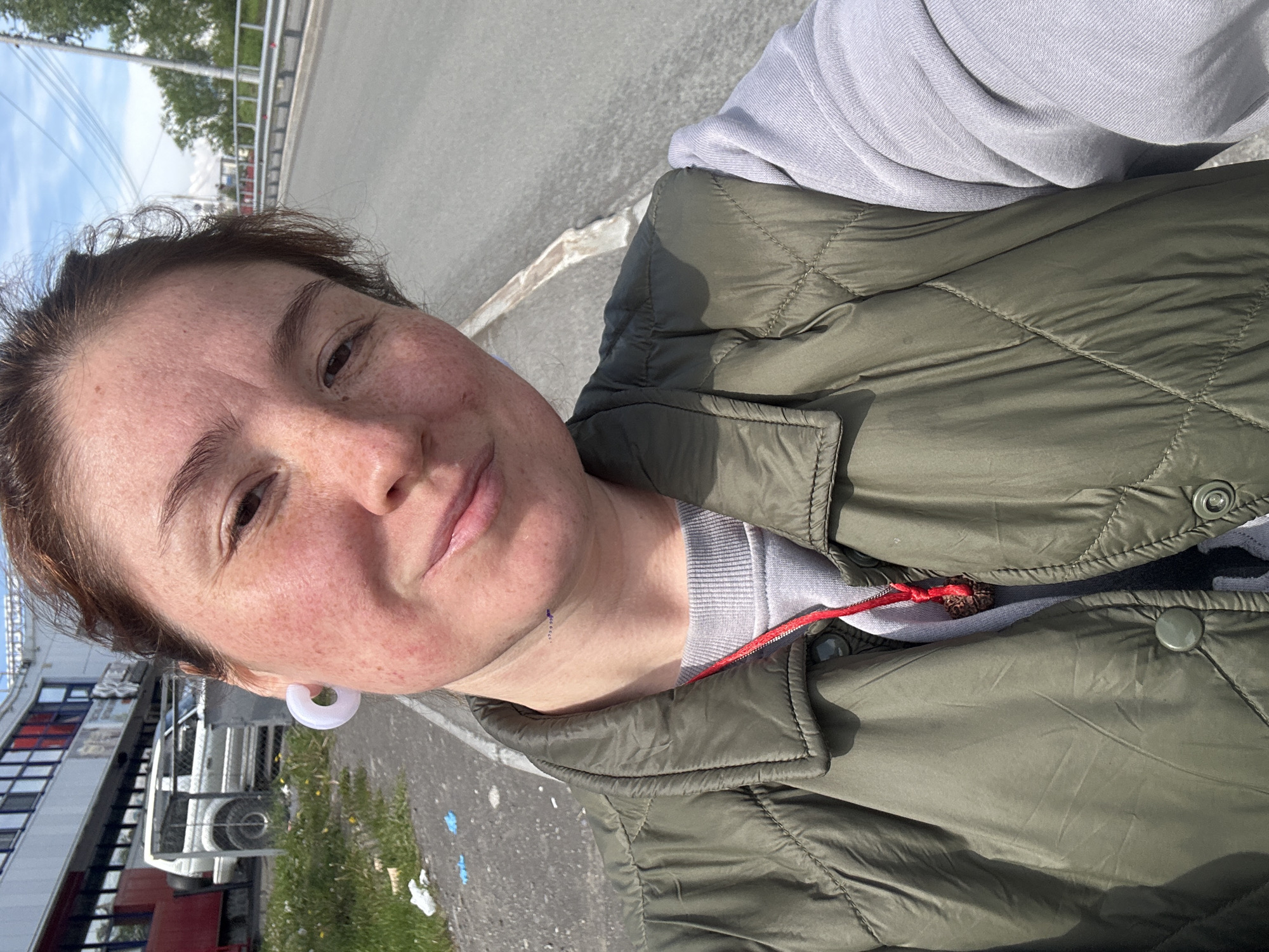 The image is a selfie taken by a woman in Petropavlovsk-Kamchatsky, Russia. She is standing outdoors, with a road and a building in the background. The woman has light skin, brown hair, and is wearing a green quilted vest over a grey sweatshirt. She has a white hoop earring in her left ear. There is trash on the ground to the left of the woman. In the background, there is a white SUV parked in front of a building with red accents. A guardrail and trees are visible further in the distance, along with a cloudy sky.