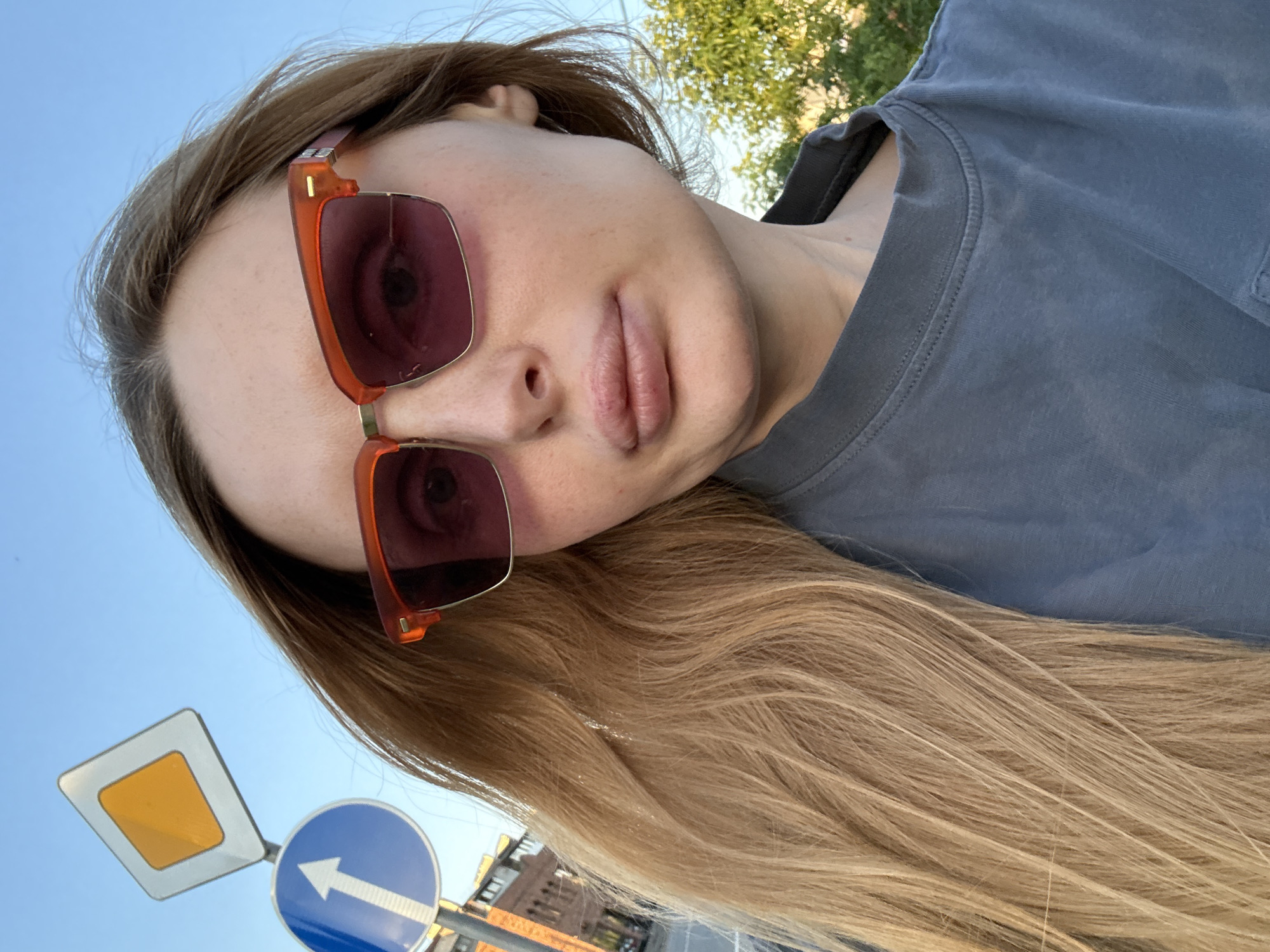 The image is a selfie taken outdoors. The main subject is a young woman with long, blonde hair, wearing a grey t-shirt and large, orange-framed sunglasses with pink lenses. Her face is partially visible, with her lips slightly parted. The background shows a clear blue sky and some green foliage, suggesting it is daytime and likely pleasant weather.
In the foreground, two road signs are visible. One is a white diamond-shaped sign with a yellow square in the center, commonly indicating a priority road. The other is a circular blue sign with a white arrow pointing to the left, indicating a mandatory direction. The signs and the woman's hair and clothing are positioned as if she is looking down or the photo is taken from a low angle, possibly while walking or standing on a street. The overall tone is casual and relaxed. There is no visible text in the image.