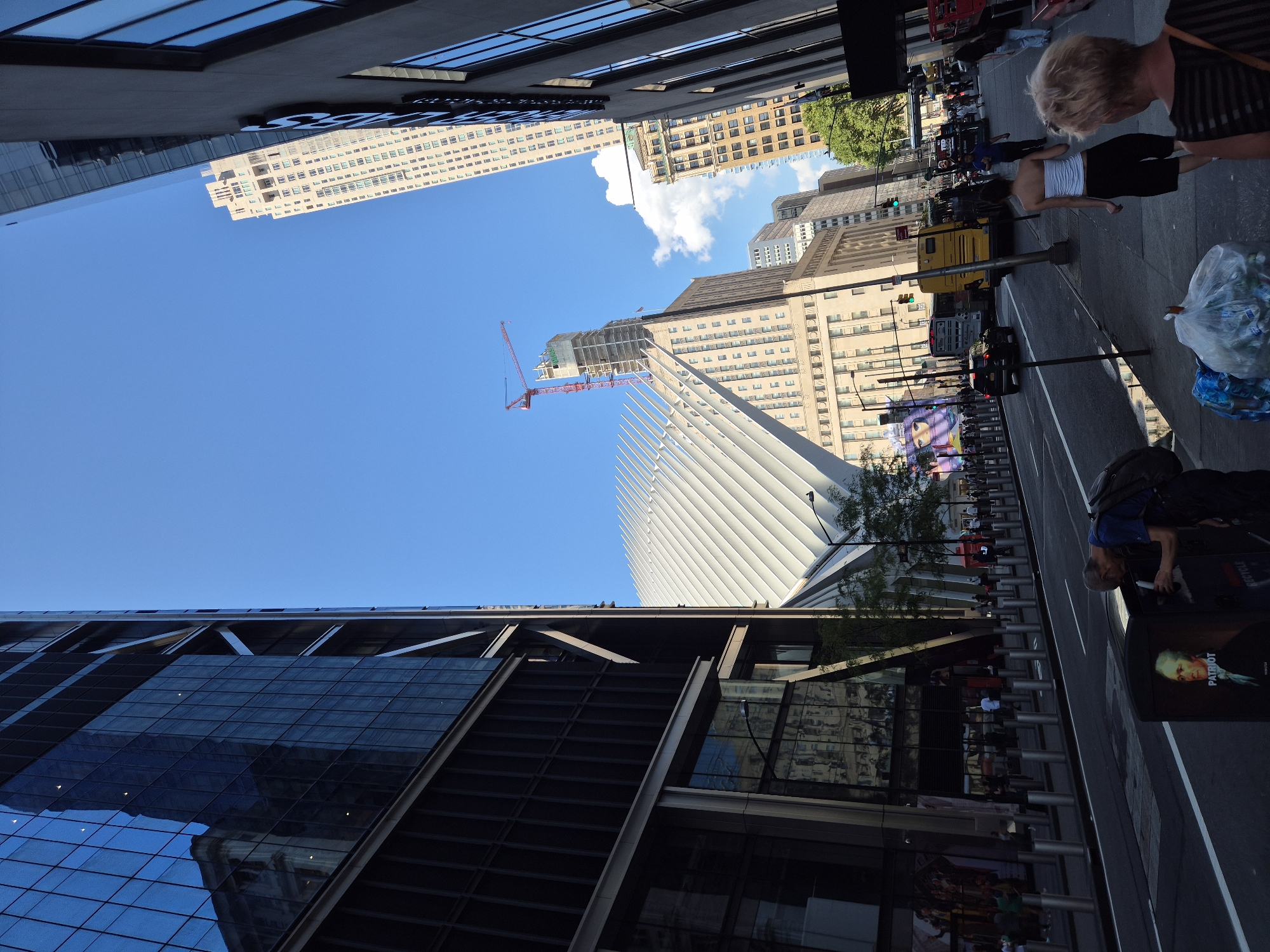 This image captures a street scene in New York City, United States of America, featuring the Oculus transportation hub and surrounding skyscrapers. In the foreground, two individuals, a woman with blonde hair and another woman in a white top and black shorts, walk on the sidewalk. A man in a blue shirt and backpack is bending down near a trash can adorned with an image of Alexander Hamilton. Across the street, a yellow bus and other vehicles are visible, with people walking on the sidewalk in the distance. The Oculus, with its distinctive white, wing-like structure, dominates the middle ground, and a construction crane is visible above a building to its left. The scene is set under a clear blue sky with a few scattered clouds, suggesting a daytime setting. The architecture suggests a modern urban environment with a mix of contemporary and older buildings.