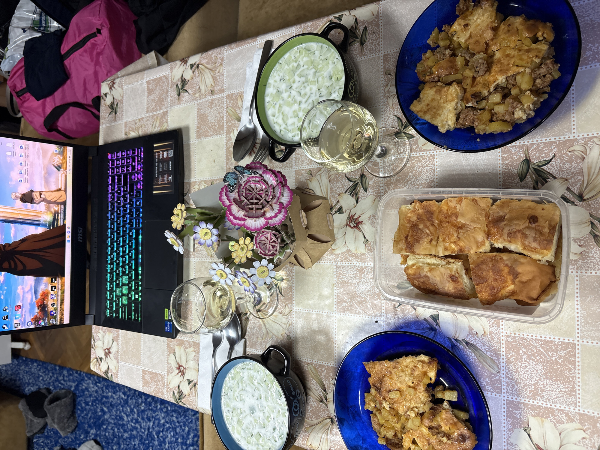 A table is set with a laptop, food, and drinks. The laptop, open and displaying an animated scene, sits on the left side of the table. Several dishes of food, including what appears to be salad in a dark-colored bowl, potato-based dishes in two blue bowls, and pastries in a plastic container, are arranged across the table. Two glasses of white wine are also present. A small bouquet of flowers decorates the center of the table. The table is covered with a floral-patterned tablecloth. The setting is Gorna Oryahovitsa, Bulgaria.