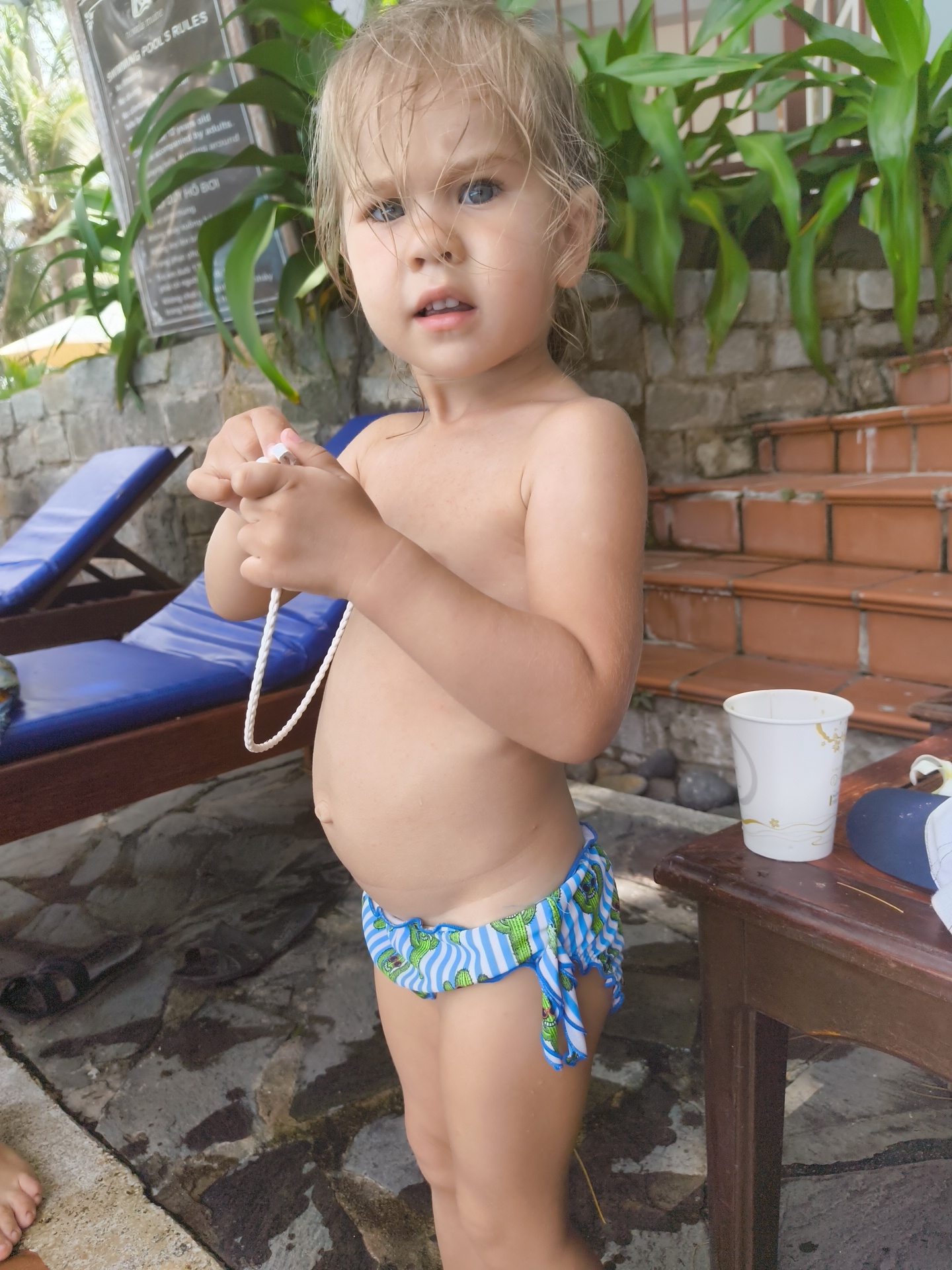 This media file is a portrait-oriented, full-body shot of a young child, likely a toddler, standing outdoors. The child has light blonde hair, which appears damp, and blue eyes. They are wearing patterned swim trunks with a blue and white striped design featuring green cacti. The child is holding a white braided cord in their hands, which hangs down in front of them. Their expression is one of slight curiosity or slight discomfort, with their mouth open and eyes looking forward.
The setting appears to be a poolside or resort area. In the background, there are two blue lounge chairs, a stone wall, and lush green foliage. To the right of the child, there are steps made of reddish-brown brick leading upwards. On a wooden table, there is a white paper cup with a decorative pattern and a blue cap. The ground beneath the child is wet and appears to be made of stone or concrete.
The lighting suggests it is daytime, with sunlight filtering through the leaves, creating dappled shadows. The overall mood is casual and indicative of a holiday or warm-weather activity.
A sign is visible in the upper left corner, with text that appears to be in English and possibly another language. The visible English text includes "SWIMMING POOLS RULES". Other text is less legible due to the distance and resolution.
The location context provided is Phu Thuy Ward, Vietnam, which aligns with the tropical foliage and resort-like environment. The activity taking place is the child standing by the pool, possibly after swimming or preparing to swim.