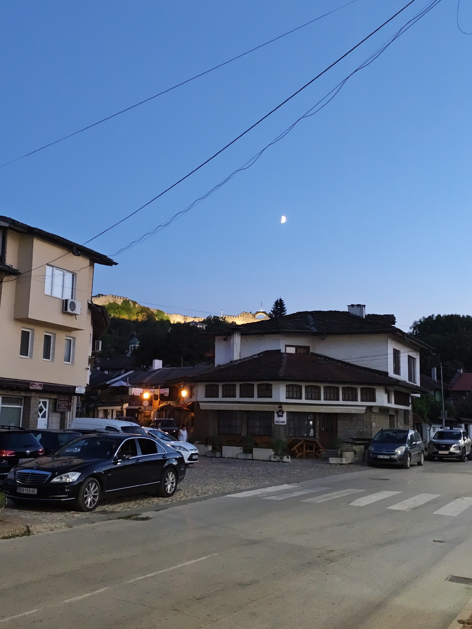 The image depicts a street scene in the city of Lovech, Bulgaria, at dusk. A black Mercedes-Benz sedan is parked on the cobblestone street in the foreground. Behind it, a white Ford is parked, followed by other vehicles. The street is lined with buildings, some of which appear to be shops or restaurants with outdoor seating. In the background, a fortress or castle is illuminated on a hilltop, overlooking the city. The sky is a deep blue, with a sliver of the moon visible. The overall impression is one of a charming, historic town transitioning from day to night.