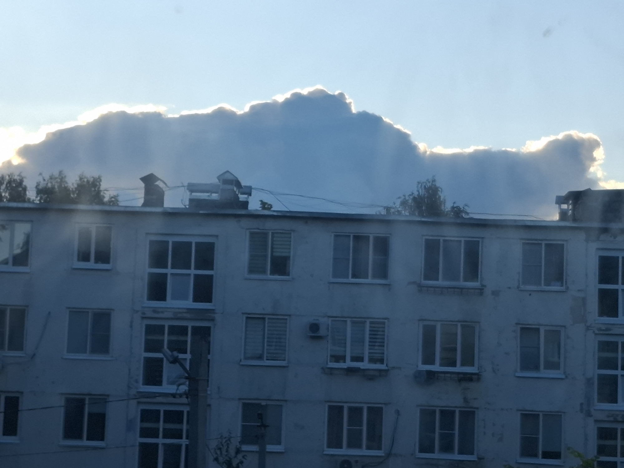 In the city of Chaikovsky, Russia, a multi-story apartment building is captured from a low angle, emphasizing the building's facade against a sky dominated by dramatic, sun-drenched clouds. The sky is a pale blue, transitioning to a bright white haze where the sun's rays pierce through the thick, cumulonimbus clouds. The clouds themselves are illuminated from behind, creating a bright, glowing halo around their edges, with shafts of light descending towards the building.
The apartment building appears to be constructed of light-colored concrete or plaster, showing signs of age and weathering with visible patches and discoloration. It is composed of multiple floors, each lined with numerous windows. These windows are mostly uniform in size and shape, with white frames, and many are divided into smaller panes. Some windows have blinds or curtains visible inside.
On the roof of the building, several chimneys and ventilation pipes are present, along with some electrical wiring. In the foreground, a utility pole with a street light fixture and some wires extends upwards along the side of the building. Trees with sparse foliage are visible along the rooftopline and peeking out from behind the building, suggesting a residential area. The overall impression is a mundane urban scene elevated by the striking natural phenomenon of the sun breaking through the clouds. There are no people or discernible actions depicted in the image.