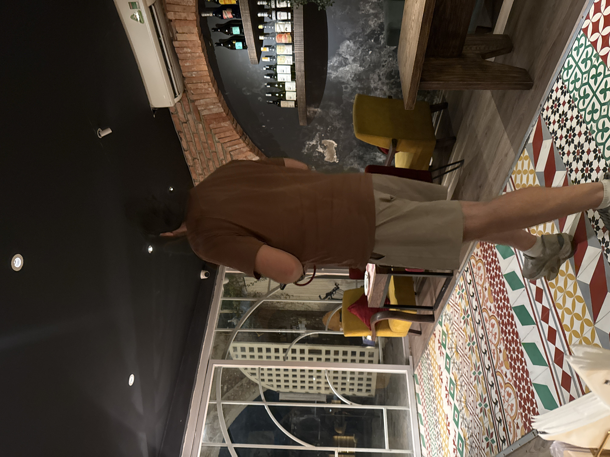 The image depicts the interior of a restaurant or bar in Hanoi, Vietnam. A man wearing a brown t-shirt and beige shorts is standing with his back to the camera, facing towards a large glass door and window with a white ornate frame. He is holding a red cord in his right hand. The room has a dark ceiling with recessed lighting and an air conditioning unit mounted on a brick arch. Shelves filled with bottles are visible on a textured, dark wall. In the foreground, a man is walking on a floor covered with a colorful, geometric patterned tile. Two yellow chairs with red cushions are visible near a wooden table. The overall ambiance suggests a modern, stylish establishment.