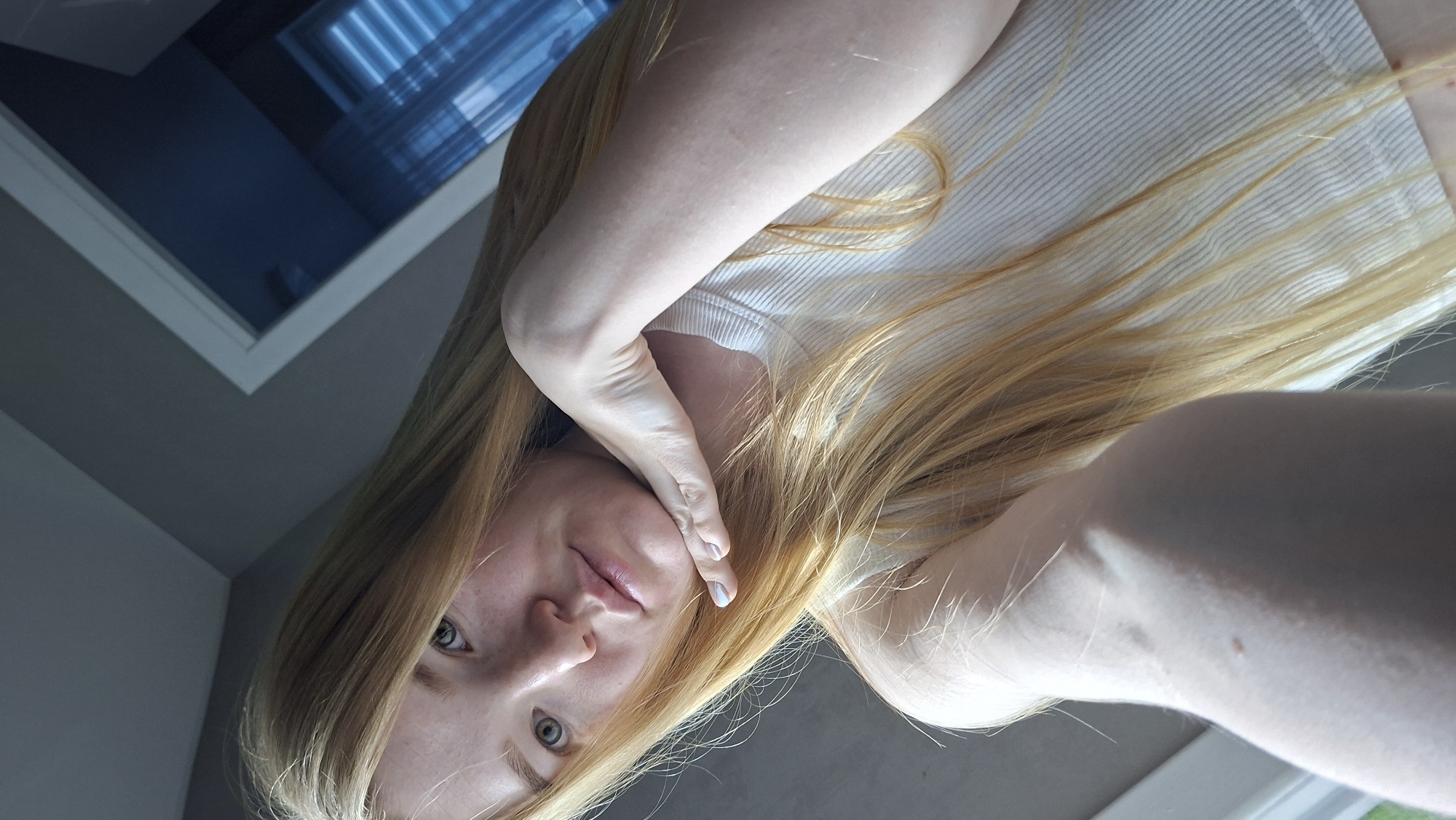 A selfie of a fair-skinned person with long, blonde hair. They are wearing a white, ribbed shirt and touching their face with their left hand. A window is visible in the upper left corner. The location is unknown, and there is no evidence to suggest that this image was taken in Chaikovsky, Russia.