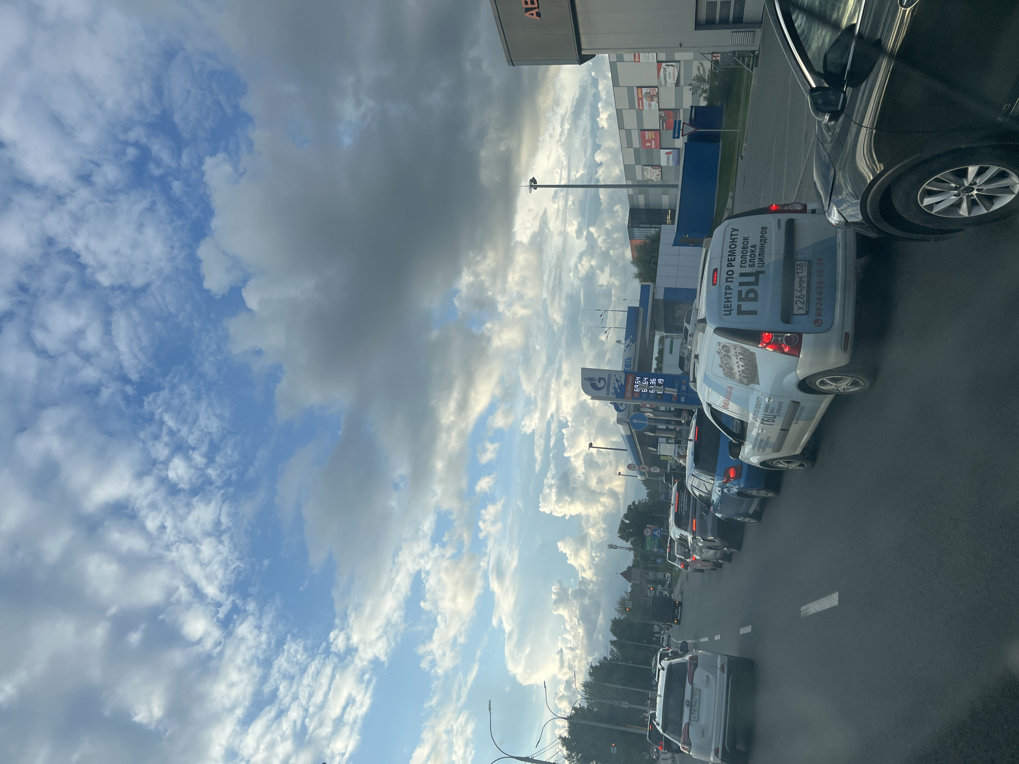The image captures a traffic jam on a city street, likely in Novosibirsk, Russia, given the Cyrillic script visible on the license plates and signage. The sky above is filled with dramatic, large white and grey clouds against a bright blue backdrop, suggesting dynamic weather conditions.
In the foreground, a black SUV is stopped in traffic, its right side mirror prominently visible. Behind it, a light-colored van with Cyrillic lettering reading "ЦЕНТР ПО РЕМОНТУ ГБЦ" (Center for Cylinder Head Repair) and a license plate starting with "Х264ММ" is also stationary. Further ahead, several other vehicles, including sedans and SUVs, are lined up on the asphalt road, which has white lane markings.
To the right of the road, a gas station is visible, featuring prominent blue and white signage with "ГАЗПРОМНЕФТЬ" (Gazpromneft) and fuel prices displayed digitally. The gas station canopy is also evident. Adjacent to the gas station, a modern-looking building with a grey and white facade, possibly a retail or commercial establishment, stands with various advertisements affixed to its exterior.
On the left side of the road, trees line the street, and streetlights are present. In the distance, buildings and structures indicative of an urban environment can be seen. The overall scene depicts a typical urban traffic scenario under a striking sky.