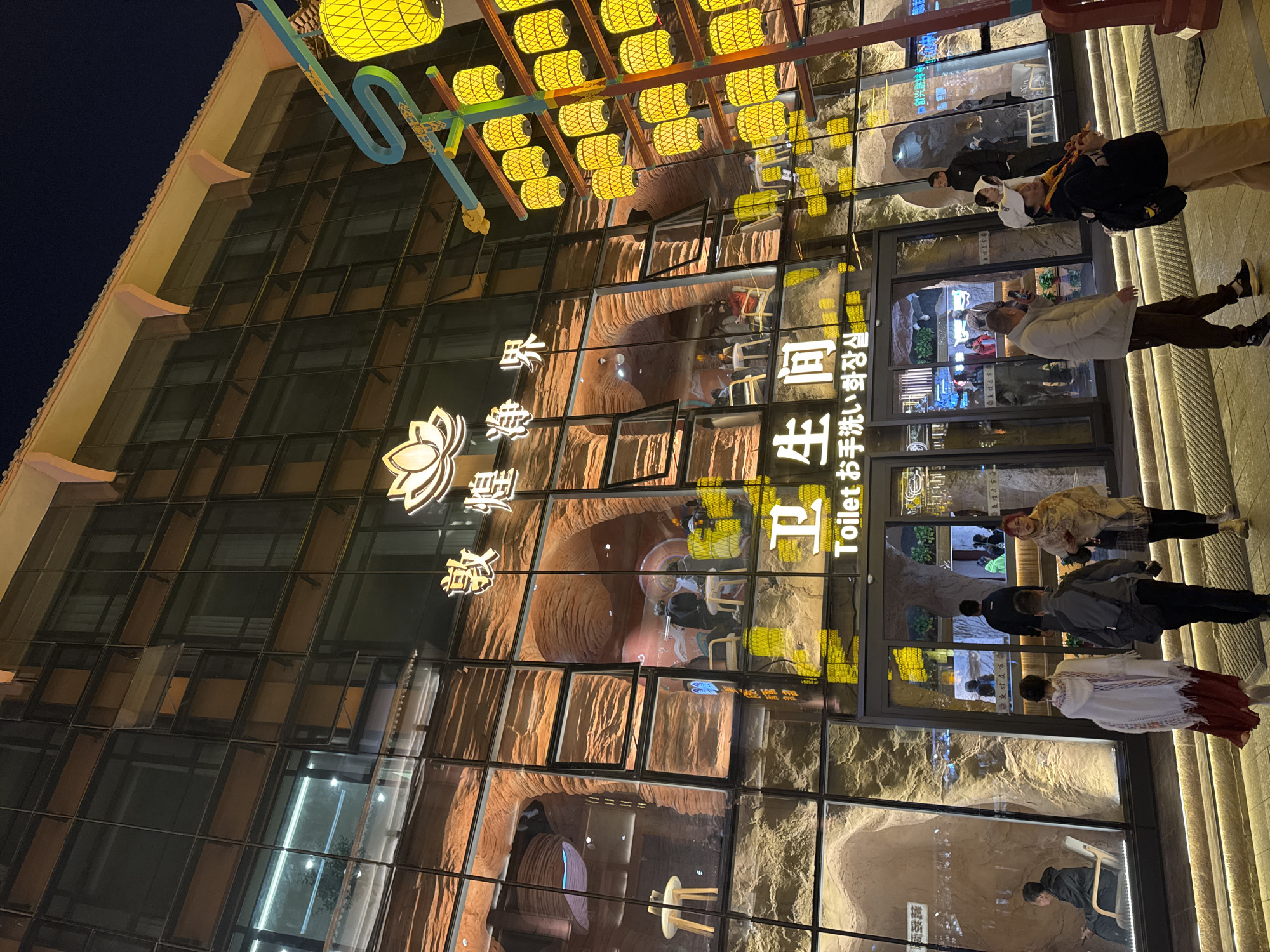 The image shows the exterior of a building in Dunhuang City, China, at night. The building has a modern glass facade reflecting an interior that evokes a cave-like environment with artificial rock formations. People are seen walking along the sidewalk in front of the building. Above the entrance, decorative yellow lanterns are suspended from a blue frame. The building's signage includes both Chinese and English text; "Toilet お手洗い洗顔".