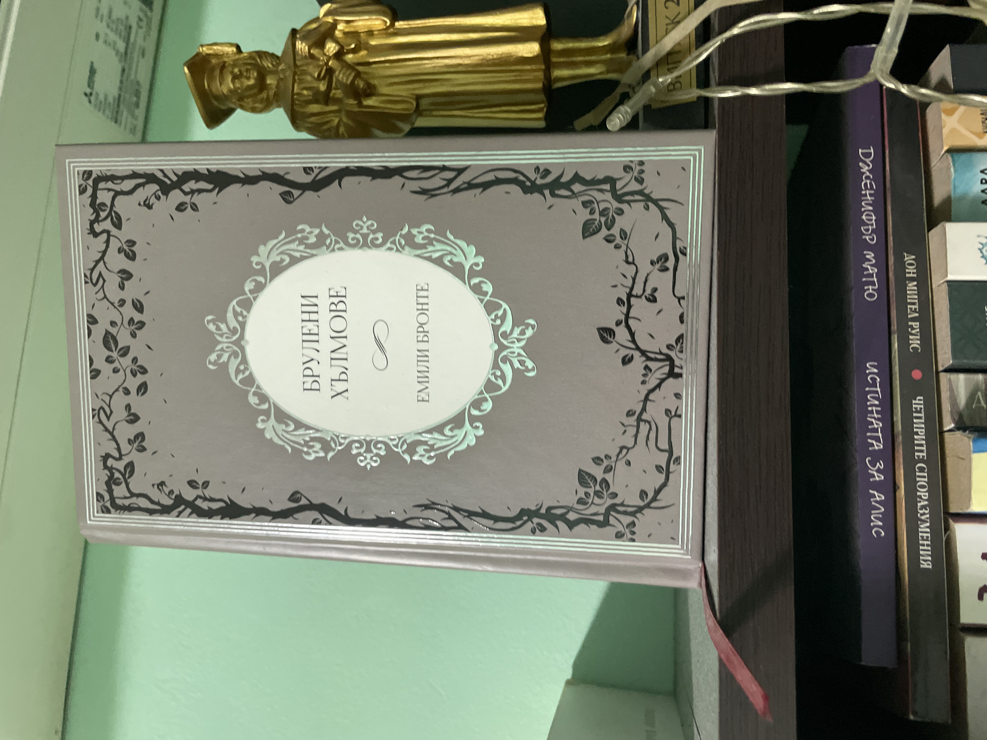 In a room with light green walls and dark wooden shelves, a closed book with a grey cover and an ornate silver design is displayed prominently. The book's title, in Cyrillic, reads "БРУЛЕНИ ХЪЛМОВЕ" (Brulyani Hylmove), and below it, "ЕМИЛИ БРОНТЕ" (Emily Bronte). To the right of the book stands a gold-colored statue of a robed figure wearing a graduation cap, holding a scroll. Below the main shelf, other books are visible, including one with a dark purple cover and Cyrillic text, and another with a dark grey cover and Cyrillic text. A red ribbon bookmark extends from the main book, and a string of white lights is draped across the lower right portion of the scene.