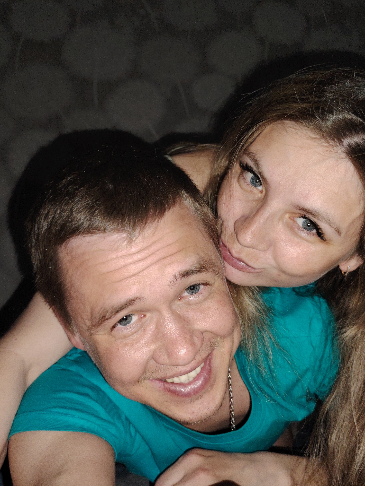 Two individuals, a man and a woman, are captured in a close-up, selfie-style photograph, conveying a sense of intimacy and happiness.
In the foreground, a man with short, light brown hair and bright blue eyes smiles broadly at the camera, revealing his white teeth. He has light stubble and a faint mustache. He wears a vibrant teal or turquoise t-shirt, and a silver chain is visible around his neck. His expression is joyful and engaging.
Behind and slightly to his right, a woman with long, flowing light brown hair and striking blue eyes rests her head on his shoulder or back. She looks directly at the camera with a soft, playful smile or pout. Her eyes are accentuated with dark eyeliner or mascara, and a small earring is visible in her right ear. She appears to be wearing a top of a similar teal or turquoise color.
Their close proximity and happy expressions suggest an affectionate relationship, possibly a couple or close friends sharing a moment. The background is dark and indistinct, appearing to be an indoor setting with a subtly textured wall. The lighting is soft and somewhat dim, creating a cozy atmosphere. There is no visible text in the image. While the location context is Chaikovsky, Russia, no specific visual cues within the photograph indicate this.