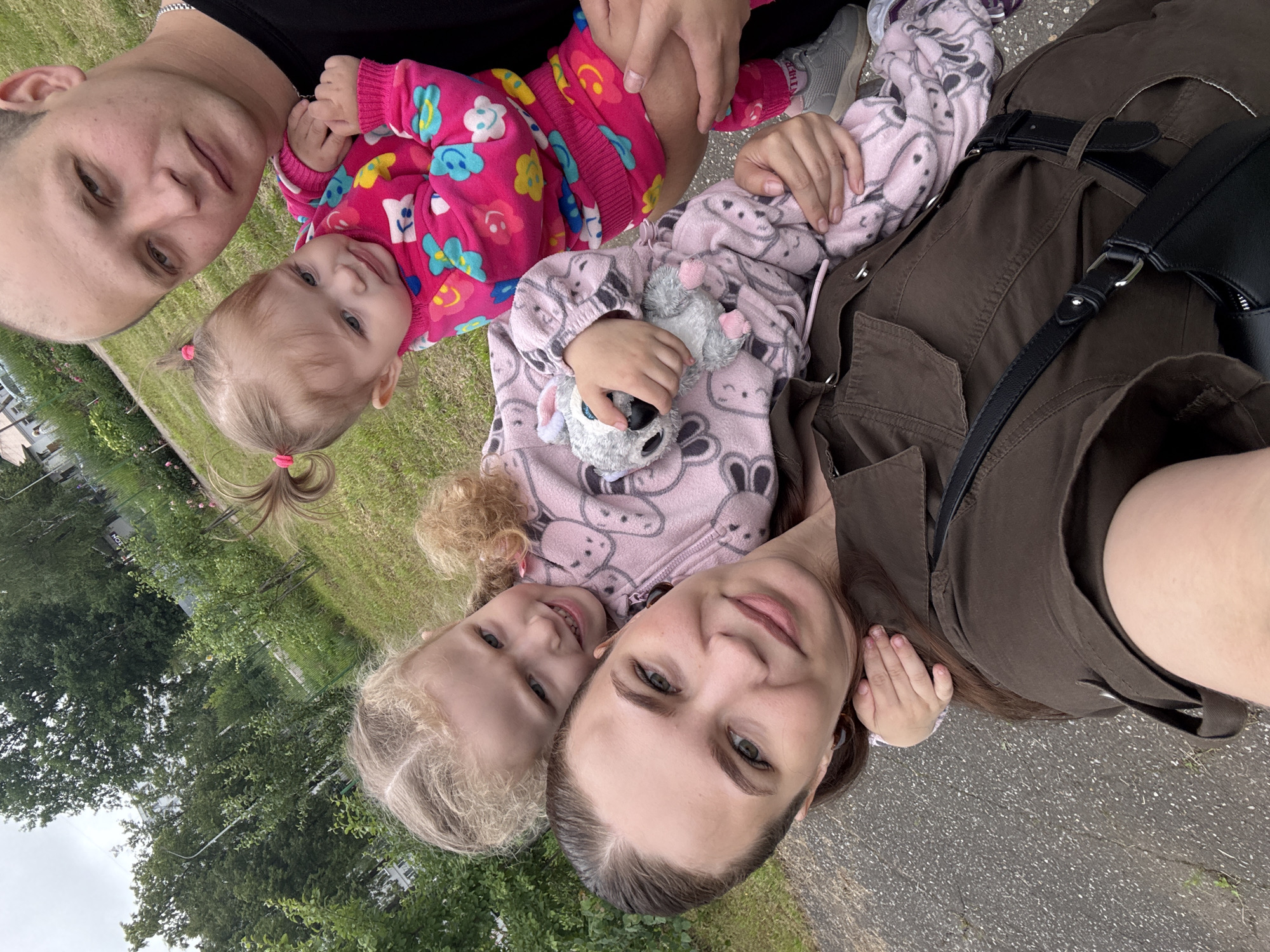 In Chaikovsky, Russia, a family of four poses for a selfie outdoors. A woman in a brown jacket is in the foreground, smiling at the camera. Behind her, a young girl with blonde hair wears a pink outfit with cartoon characters and holds a stuffed animal. To the right, a baby in a pink outfit with floral patterns is held by a man with a shaved head wearing a black shirt. The background consists of a grassy area with trees and a building in the distance. The scene appears to be a casual, everyday moment captured on an overcast day.