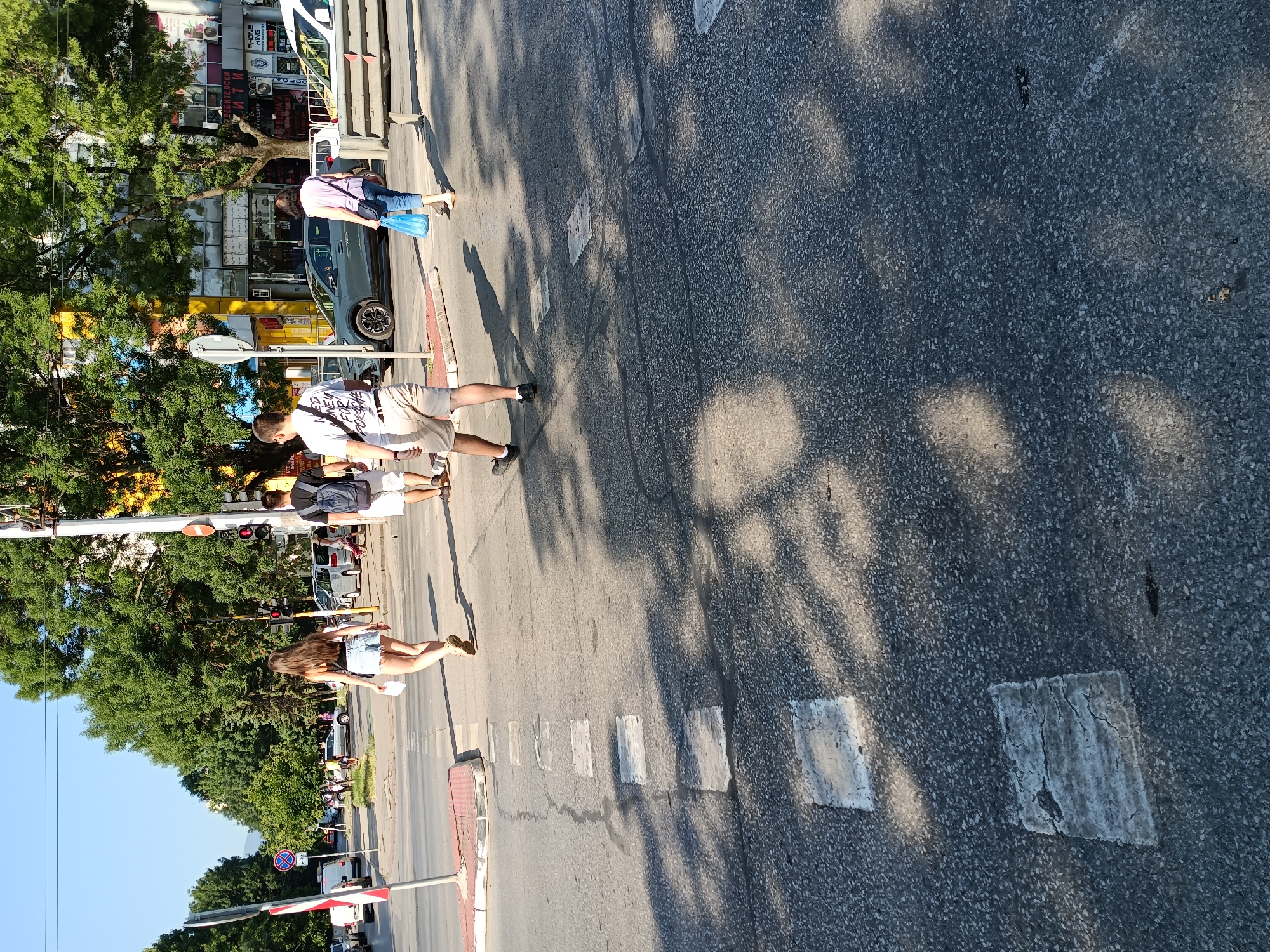 Four pedestrians are crossing a street at a marked crosswalk under a clear sky. The scene appears to be in Sofia, Bulgaria, indicated by the architecture, signage, and the presence of a car with Bulgarian license plates. The time of day is likely afternoon, as evidenced by the long shadows cast by the trees and pedestrians.
In the foreground, two individuals are walking away from the camera towards the right. The person on the left is a young woman wearing a black tank top and denim shorts. She is holding a white piece of paper in her left hand and her right hand is by her side. The person on the right is a man wearing a white t-shirt with a graphic print and light-colored shorts. He has a backpack on his back.
Further ahead, two more pedestrians are crossing from left to right. The person on the left is a man wearing a dark t-shirt and white shorts, also with a backpack. The person on the right is a woman wearing a pink top and blue jeans, carrying a light blue plastic bag.
The street is asphalt, with painted white lines marking the crosswalk. There are several vehicles visible in the background, including cars and a white van. Trees line the sidewalk, and buildings with shops are visible behind them. Traffic lights and street signs are also present. The overall impression is a busy urban intersection on a sunny day.