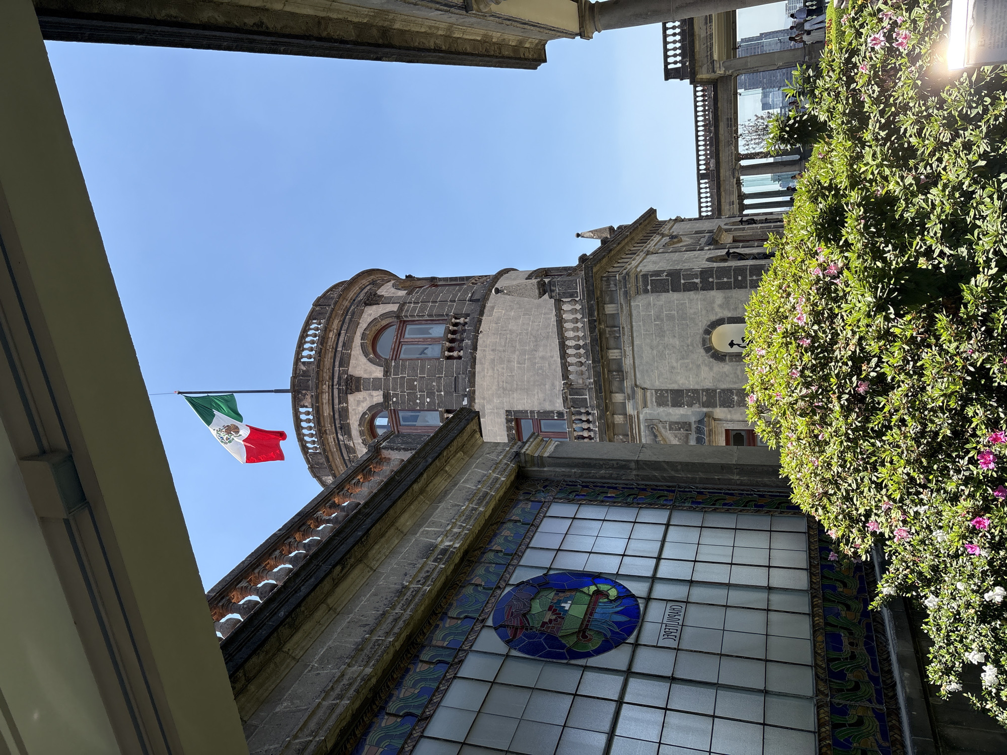 The image depicts a view of the Castillo de Chapultepec in Mexico City, Mexico, captured from a low angle. The Mexican flag is visible atop the castle. The castle's architecture is characterized by stone construction, a rounded tower, and intricate detailing. A large window with a decorative, circular design is in the foreground. Lush green vegetation with flowers frames the right side of the view. In the background, a modern glass building is visible, standing in contrast to the historical structure. The sky is clear and blue. Some people are on the top balcony of the building.