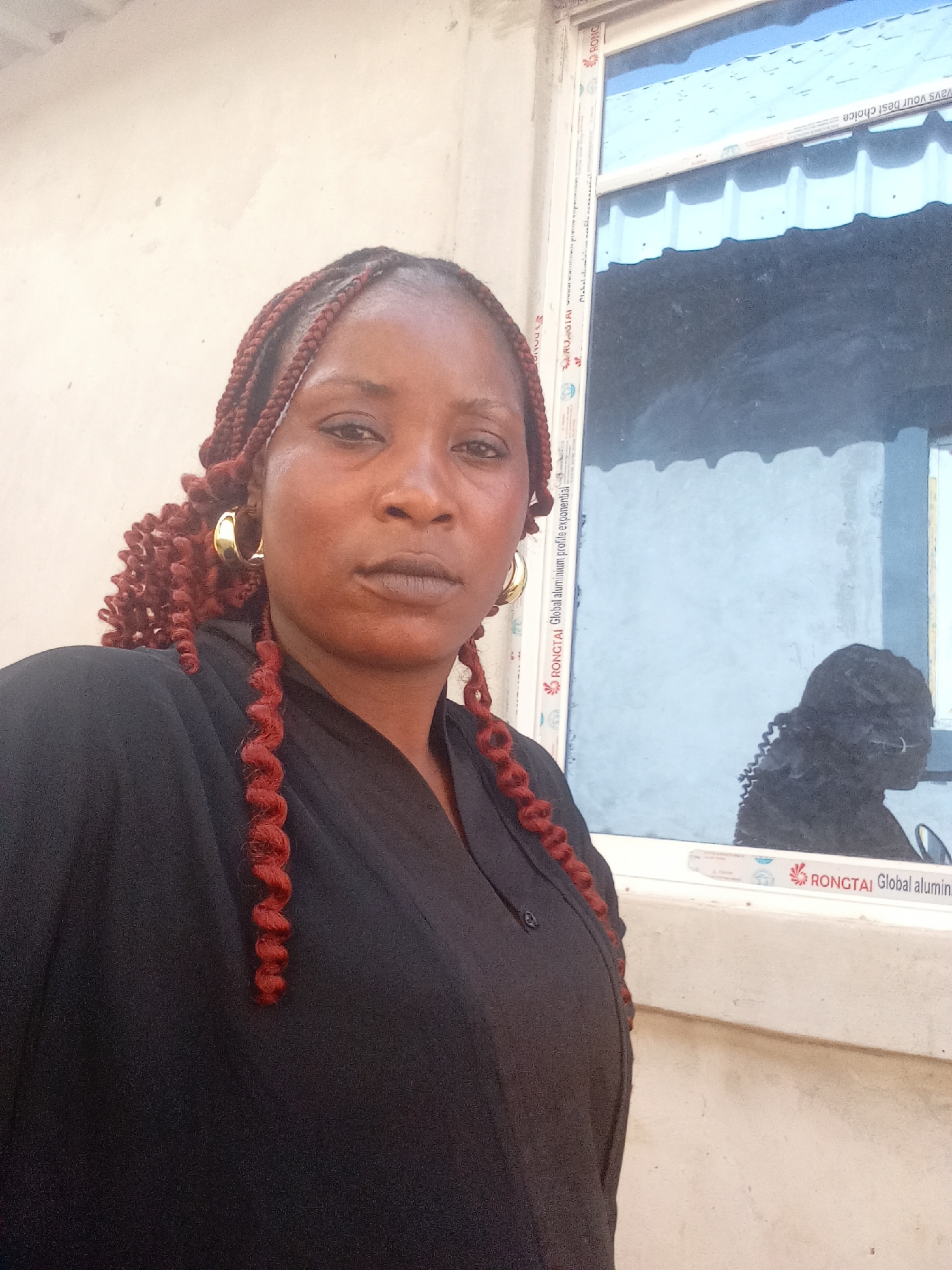 The image captures a woman in what appears to be an outdoor setting in Doma, Nigeria. She stands facing the camera, wearing a dark-colored top and large gold hoop earrings, with her hair styled in braids with reddish-brown curls at the ends. The background features a light-colored wall and a window with visible text on the frame, possibly indicating the brand or material of the window. A reflection can be seen in the window.