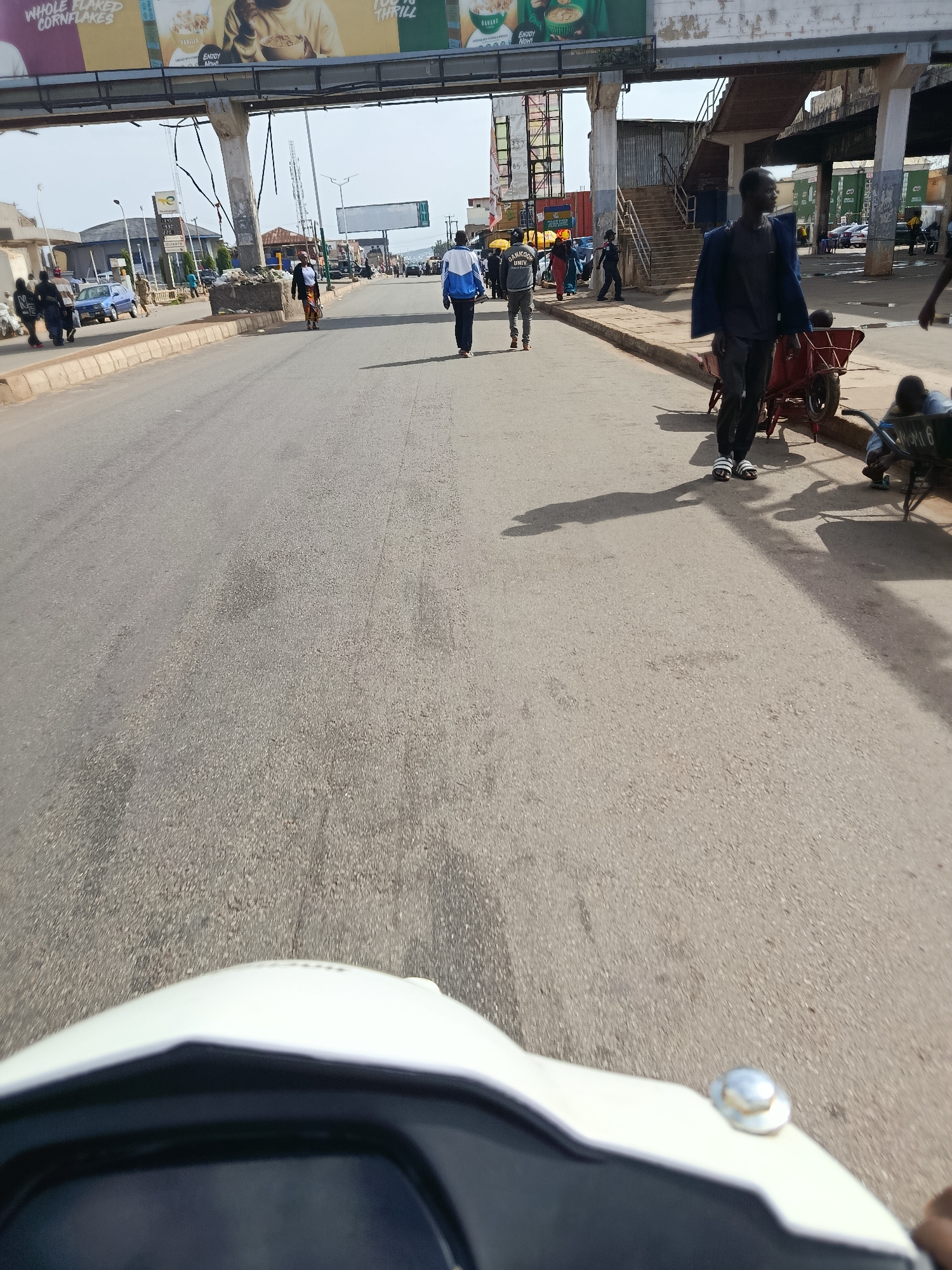A first-person perspective of a street in Jos, Nigeria, shows a road with people walking and a vehicle in the foreground. The road is paved and appears to be in a commercial or residential area. On the left side of the road, there are buildings and what looks like an overpass or bridge. Several people are walking on the sidewalk and on the road. One person in the foreground is wearing a blue and white jacket and black pants. Another person in the background is wearing a dark jacket and black pants. There are also some vehicles visible on the side of the road. The sky is overcast, and the overall lighting suggests it is daytime. In the immediate foreground, the front of a white vehicle, possibly a motorcycle or scooter, is visible, suggesting the photo was taken from a moving vehicle. The image captures a slice of everyday life in this urban environment.