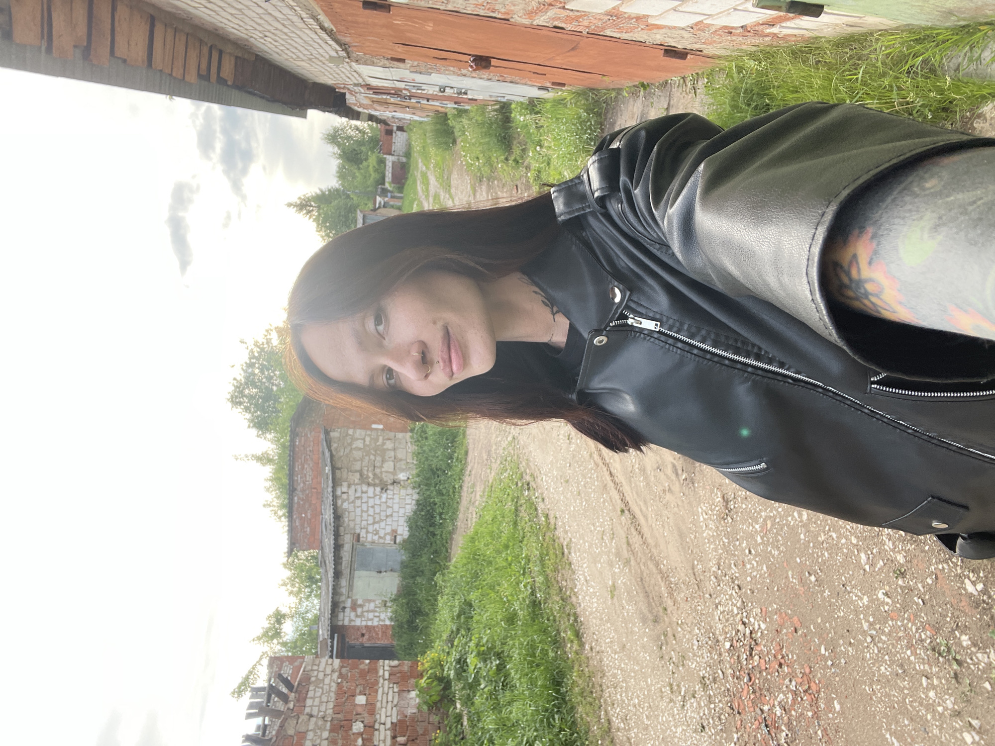 A person with long brown hair and a black leather jacket is standing on a dirt path. The person has a pierced nose, visible tattoos on their neck and arm. The background features brick buildings with closed garage doors and overgrown vegetation. The location is inferred from the metadata to be in Oktyabr'skii, Russia.