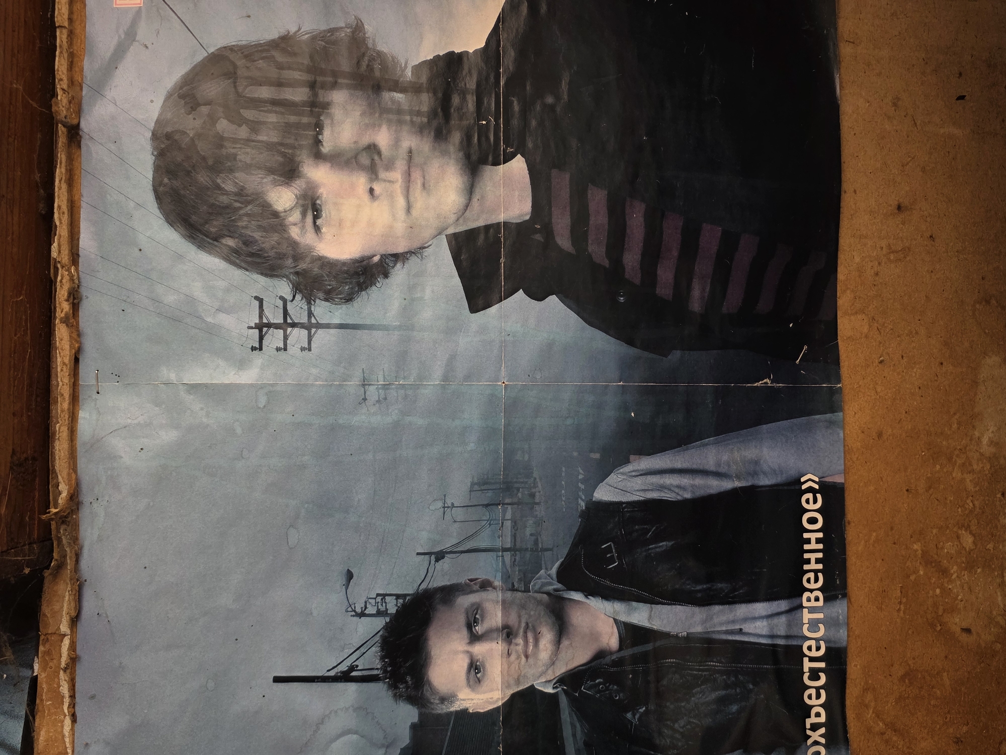 A worn, faded poster is affixed to a surface that appears to be either deteriorated wood or concrete. The poster features two individuals, one positioned above the other. The upper figure is a young man with short, dark hair, wearing a dark jacket with purple striped sleeves. The lower figure is a man with short, spiked dark hair, a gray hoodie, and a black leather jacket. In the background, visible between the figures, are utility poles and a street light. To the right of the lower figure are Russian words written vertically. The location, Зипуново, Russia, is not visually discernible from the poster alone.