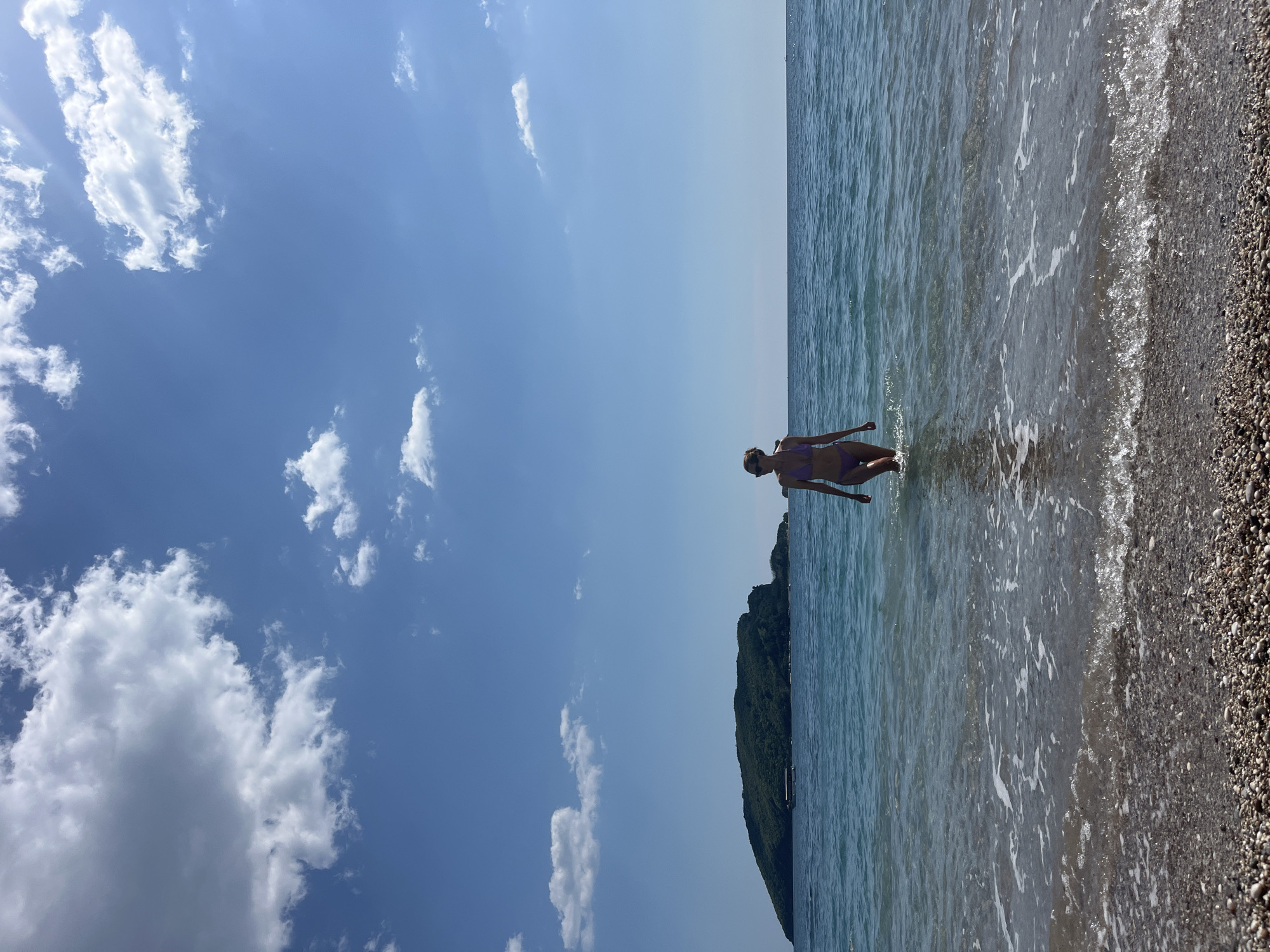 The scene is set on a beach in Budva, Montenegro, with a view of a clear blue sky dotted with white clouds. A woman is wading into the water, dressed in a bikini. The beach is pebbly, and the water is calm with small waves. In the distance, there is a forested hill or island. The scene is taken in daylight with clear visibility.