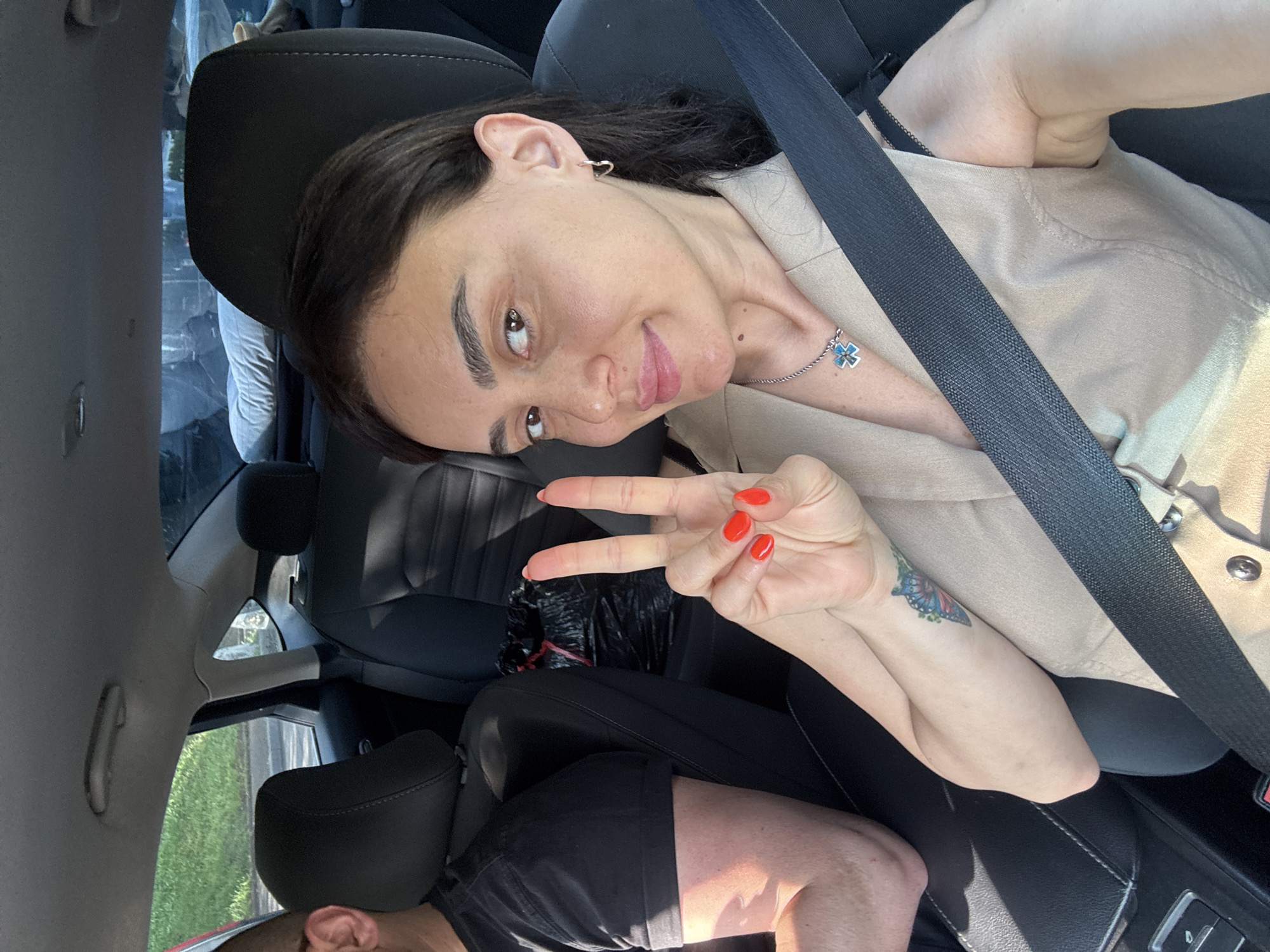 A young woman with dark hair and a tattoo on her left arm is taking a selfie in the passenger seat of a car. She is wearing a beige sleeveless top and a silver necklace with a blue cross pendant. She is holding up two fingers in a peace sign. To her left, a man with short hair wearing a black t-shirt is visible. The car appears to be in motion, as indicated by the blurred foliage and road visible through the windows. The scene takes place during daylight hours. The specific location in the city of Tbilisi, Georgia is not determinable from the image.
