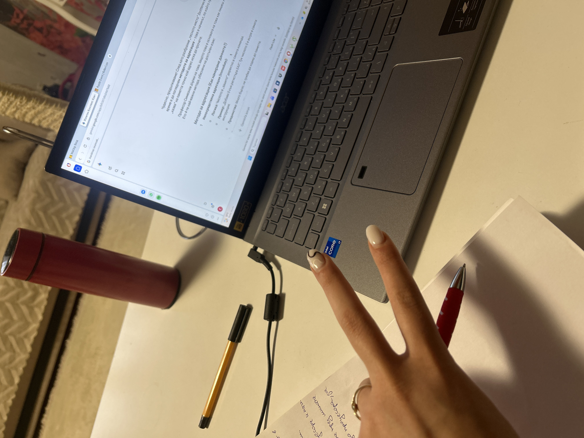 Here's a detailed description of the image:
**Scene:** The image shows a laptop sitting on a white desk. A red thermos is positioned to the left of the laptop, and a notepad with handwritten text is on the right. A hand is making a peace sign above the notepad, with a red pen resting on the notepad. A gold pen is situated near the laptop charging cable. The laptop screen displays a webpage, apparently from Gemini AI.
**Subjects:** The primary subjects are the laptop, the hand with the peace sign, the writing utensils, and the thermos.
**Objects:** Key objects present include the laptop, thermos, notepad, red pen, gold pen, and charger.
**Actions:** The hand is making a peace sign. Someone is presumably working or studying, as indicated by the laptop, notepad, and writing utensils.
**Environment:** The environment is a workspace, specifically a desk. Light seems to be indoor lighting.
The OCR text suggests that the writing on the notepad may be related to addresses. The text on the laptop screen is in Bulgarian.
There is no information in the image to suggest it is from Burgas, Bulgaria.