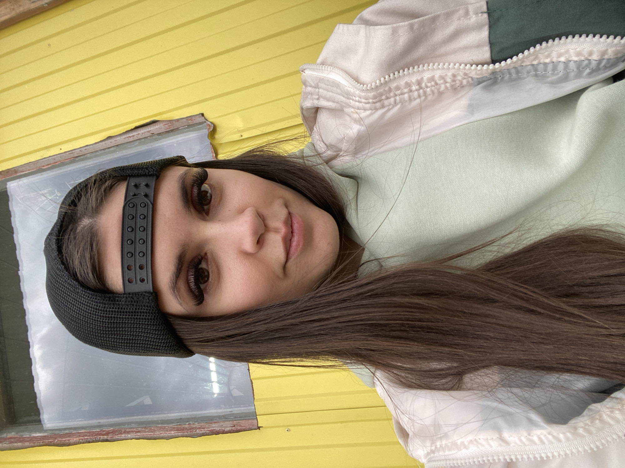 A woman with long, dark brown hair is facing the camera, wearing a black cap, a light green sweatshirt, and a white and green jacket. She is positioned in front of a yellow wall with a window to the left. The background appears to show a cloudy sky reflected in the window. The setting is Zhigalki, Russia.