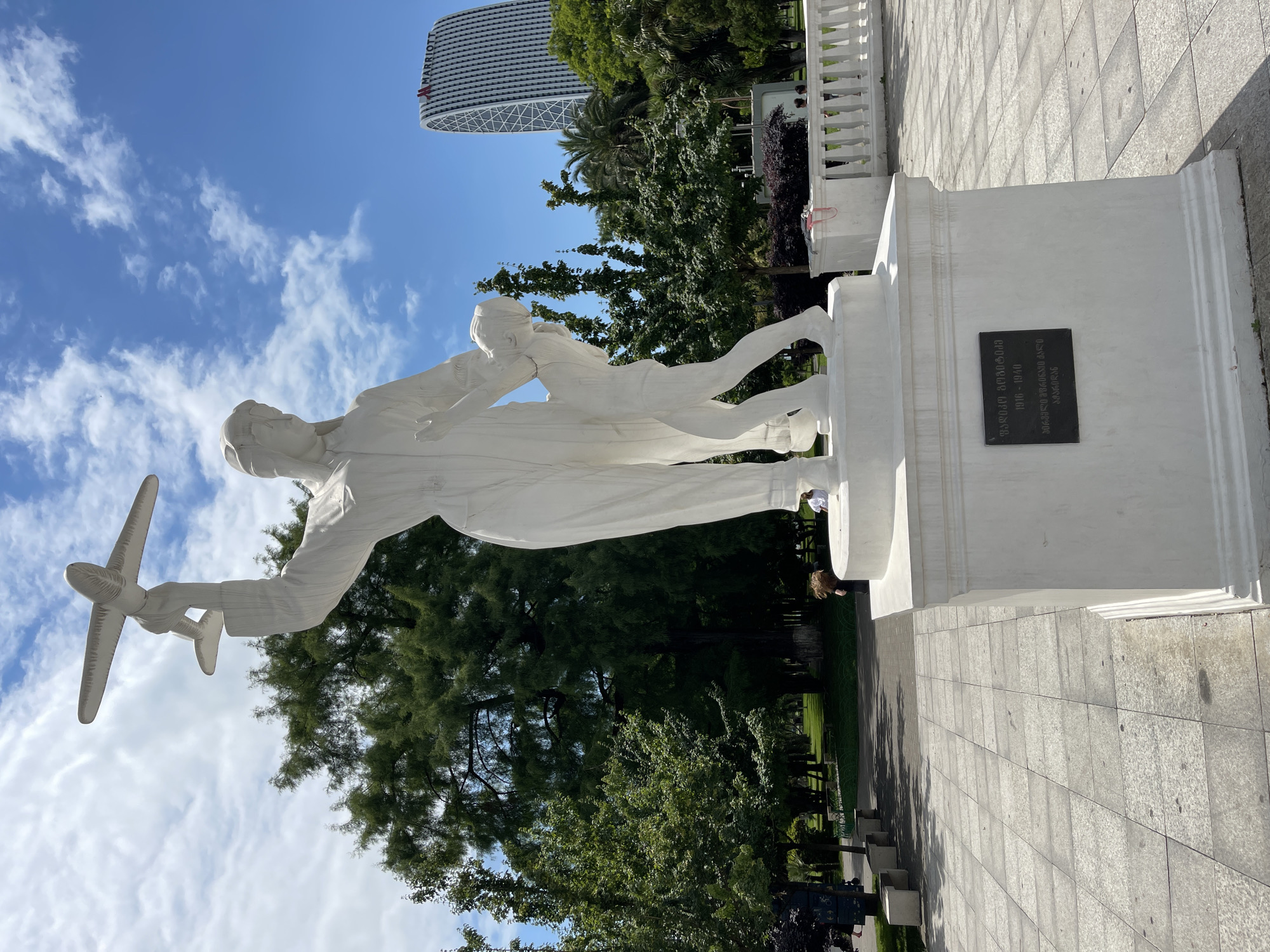 This is a vertical, eye-level, outdoor shot of a white marble sculpture depicting a pilot holding a small airplane and a child. The sculpture is on a white pedestal with a black plaque. The background features lush green trees, a modern building with a distinctive curved facade, and a clear blue sky with scattered white clouds. The scene suggests a sunny day. The plaque has text in Georgian script, which appears to be a memorial inscription. The overall impression is one of remembrance and perhaps inspiration. The composition is slightly tilted, adding a dynamic perspective to the image.
