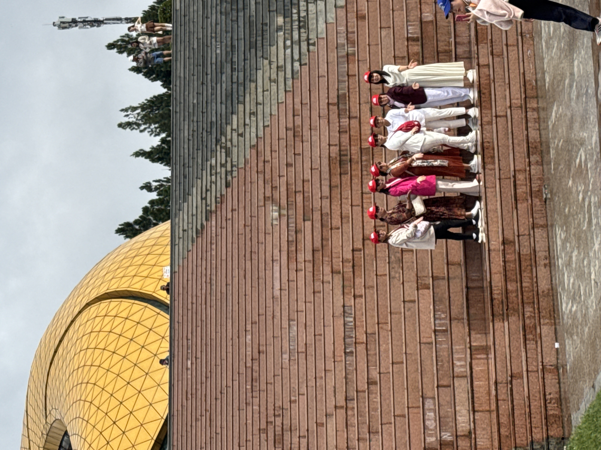 The image displays a group of people gathered on a wide, tiered platform or staircase made of reddish-brown tiles. To the left, a striking golden dome structure with a geometric, grid-like pattern dominates the midground, suggesting a modern or unique architectural landmark. The background features green trees and a tall communication tower silhouetted against an overcast sky.
A line of eleven individuals, mostly women, is positioned across several steps of the platform. They are dressed in a mix of traditional and modern attire, including long white, maroon, pink, and brown garments. Several individuals in this group are wearing matching red baseball caps, indicating they might be part of a tour group or a coordinated outing. They appear to be posing or resting during their visit.
Further to the right and slightly higher on the steps, another person, dressed in a light-colored hoodie, black pants, and a blue baseball cap, is seen holding a smartphone, seemingly capturing a photo or video. A few other indistinct figures are visible near the distant trees.
The scene depicts a daytime setting under soft, diffused light from the overcast sky, implying pleasant weather for outdoor activities. The prominent golden dome and large public steps suggest a popular tourist attraction in Da Lat, Vietnam, aligning with the provided location context. The general mood appears relaxed and social. No visible text is present in the image.