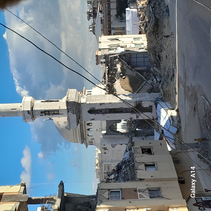 The image captures a scene of extensive urban destruction in Khan Yunis, Palestinian Territory, under a partially cloudy blue sky. Centrally positioned is a large, light-colored mosque featuring a prominent dome and a tall, multi-tiered minaret. The mosque's lower sections and surrounding structures exhibit significant damage, including visible structural compromises at the minaret's base. The building immediately to the mosque's right has largely collapsed inward, forming a substantial mound of debris.
In the foreground-left, a multi-story residential-type building is severely damaged, with large sections of its exterior walls missing, exposing interior rooms and rubble piles. Air conditioning units are affixed to its remaining facade. The ground throughout the scene, particularly in the foreground and middle ground, is covered with rubble, dirt, and sand. A relatively clear, unpaved path or street traverses the lower portion of the image.
Two individuals are present: one person wearing a light shirt and dark pants walks amidst the debris in the middle-right, and another individual in a yellow shirt is partially visible in the bottom-left, standing near a pile of rubble. White tarp-covered structures, possibly makeshift shelters, are visible among the debris piles in the middle ground. Overhead, several dark power lines or cables diagonally span the upper-middle and right sections of the frame.