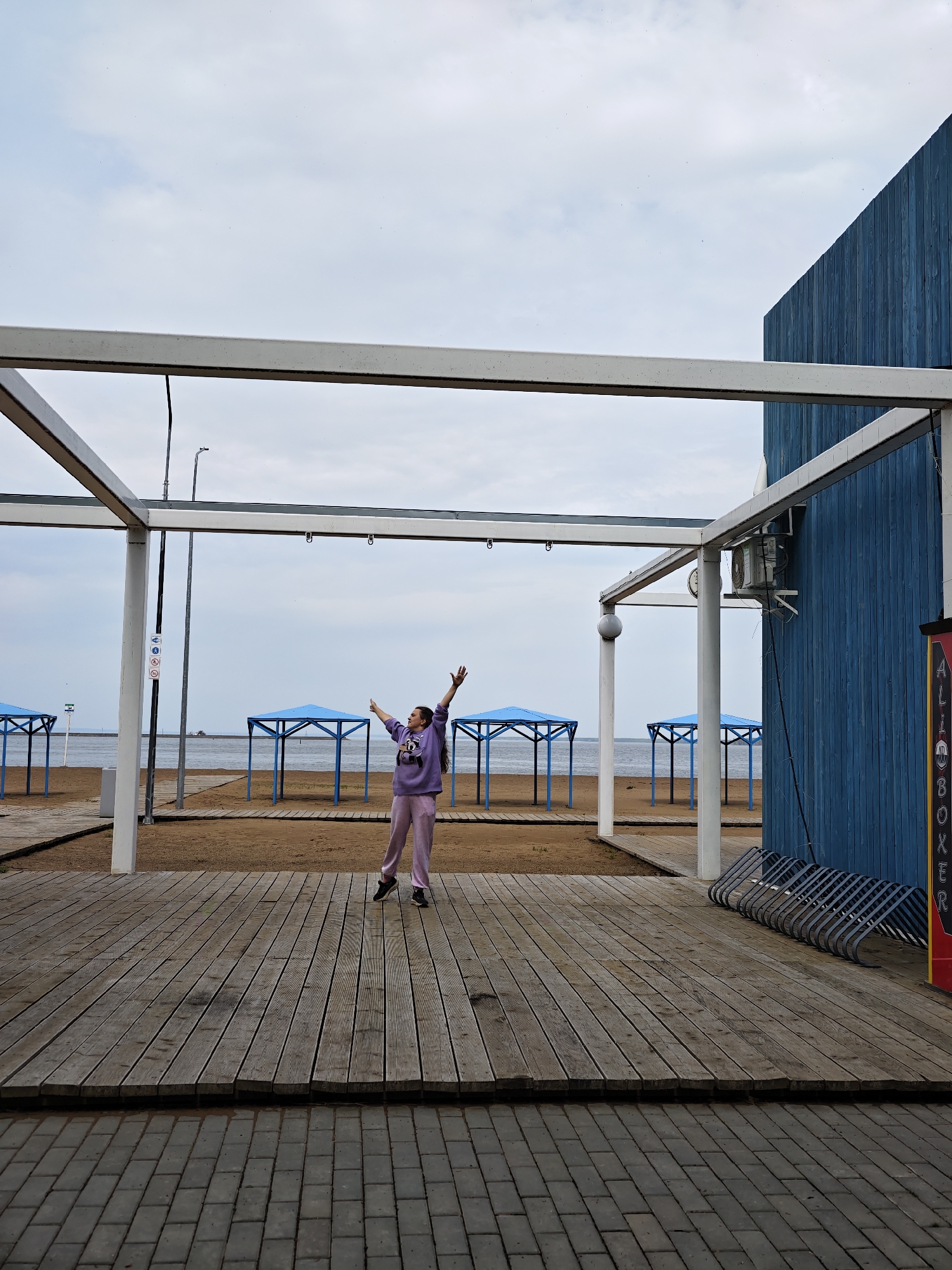 The image depicts a woman standing on a wooden platform structure at a beachfront location. She is wearing a lavender-colored sweatshirt and matching sweatpants, with her arms raised in the air. The location is identified as the city of Chaikovsky, Russia. The background shows a sandy beach with blue-roofed cabanas or sun shelters. Behind the platform, there is a blue wooden building and a bike rack. The sky is overcast. The ground in the foreground is paved with grey bricks.