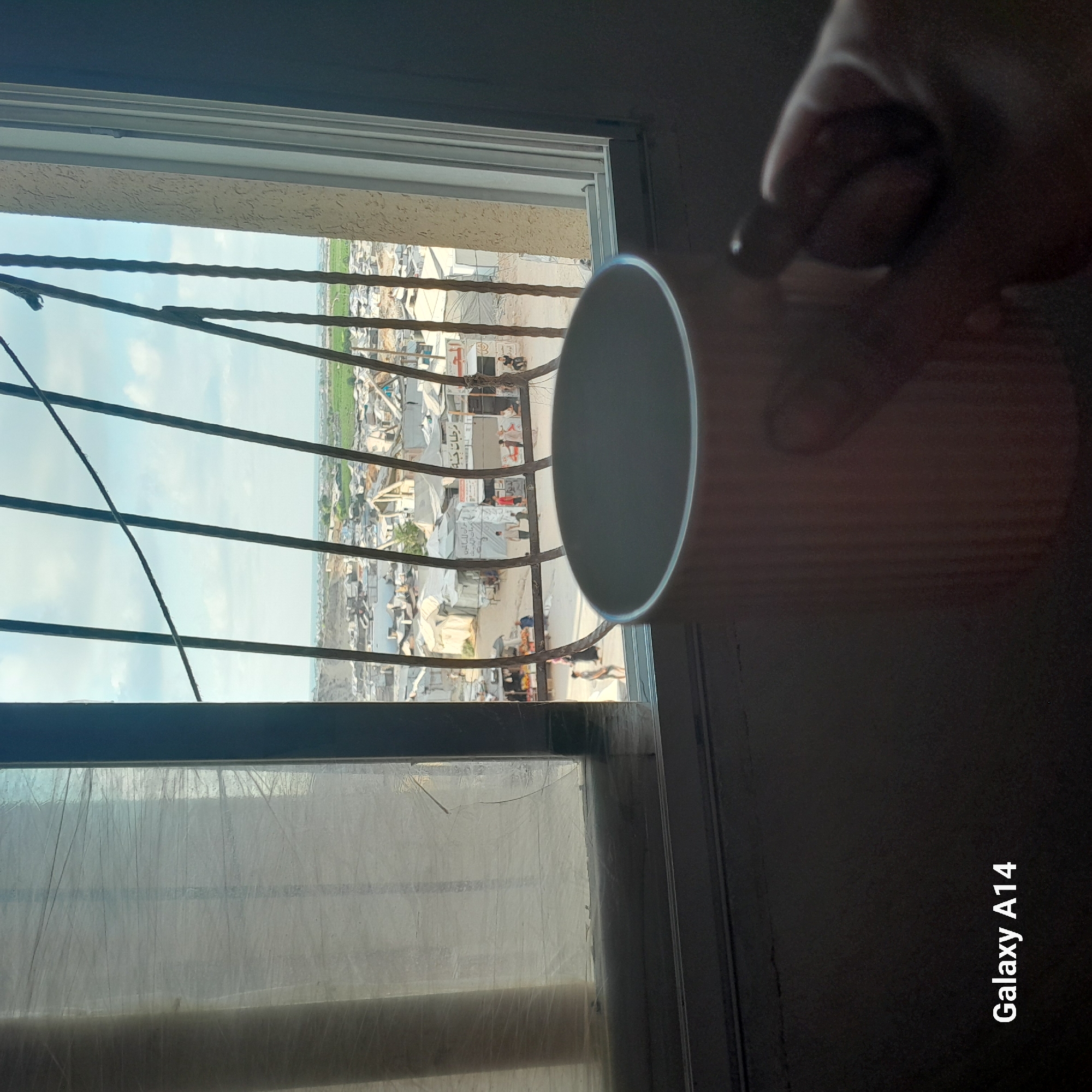 A first-person perspective shot captures a view through a barred window overlooking a densely populated area, identified as Khan Yunis, Palestinian Territories. In the foreground, a light pink ribbed mug occupies the lower right quadrant, with a portion of a hand and thumb visible on the right, holding it. The window, framed by a dark grey edge on the right and a textured beige trim, has a translucent, crinkled plastic covering on the left side. Through the bars of the window, a sprawling scene of makeshift shelters and buildings unfolds under a bright, partly cloudy sky. The shelters appear to be constructed from corrugated metal, canvas, and salvaged materials, clustered together. In the mid-ground, several individuals are visible walking and standing in what appears to be a street or open area between the structures. Signs with Arabic script are present on some of the buildings. The overall impression is of a crowded urban environment, potentially a refugee camp or a densely populated district. The "Galaxy A14" watermark is present in the lower left corner of the image.
