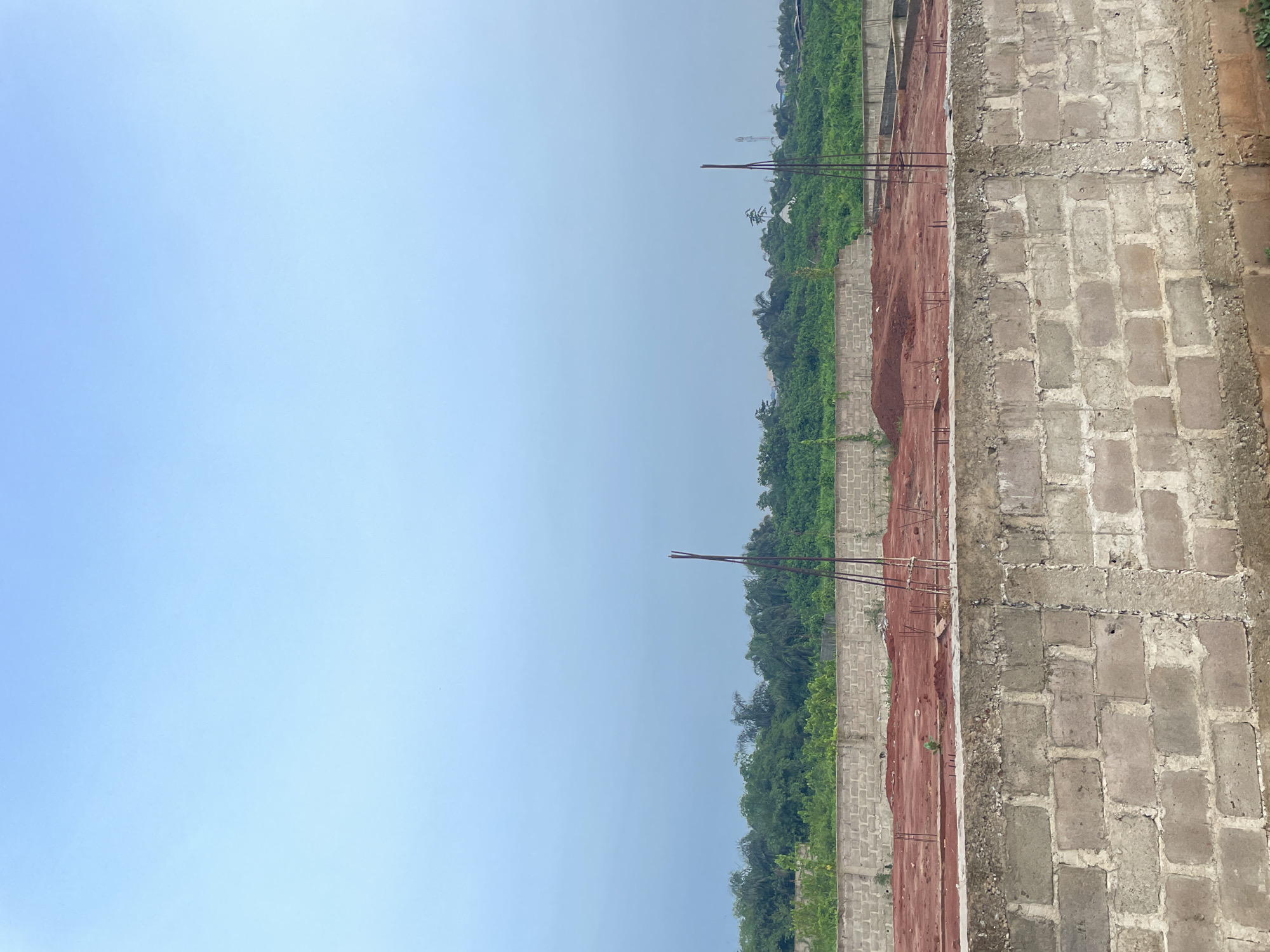 The image depicts a construction site in the city of Unknown, Unknown. A wall made of grey rectangular bricks is prominent on the right side of the image, while a pile of red earth occupies the middle ground. Green vegetation lines the background, meeting the horizon under a clear, light blue sky. Rebar sticks out from the earth, indicating ongoing construction.