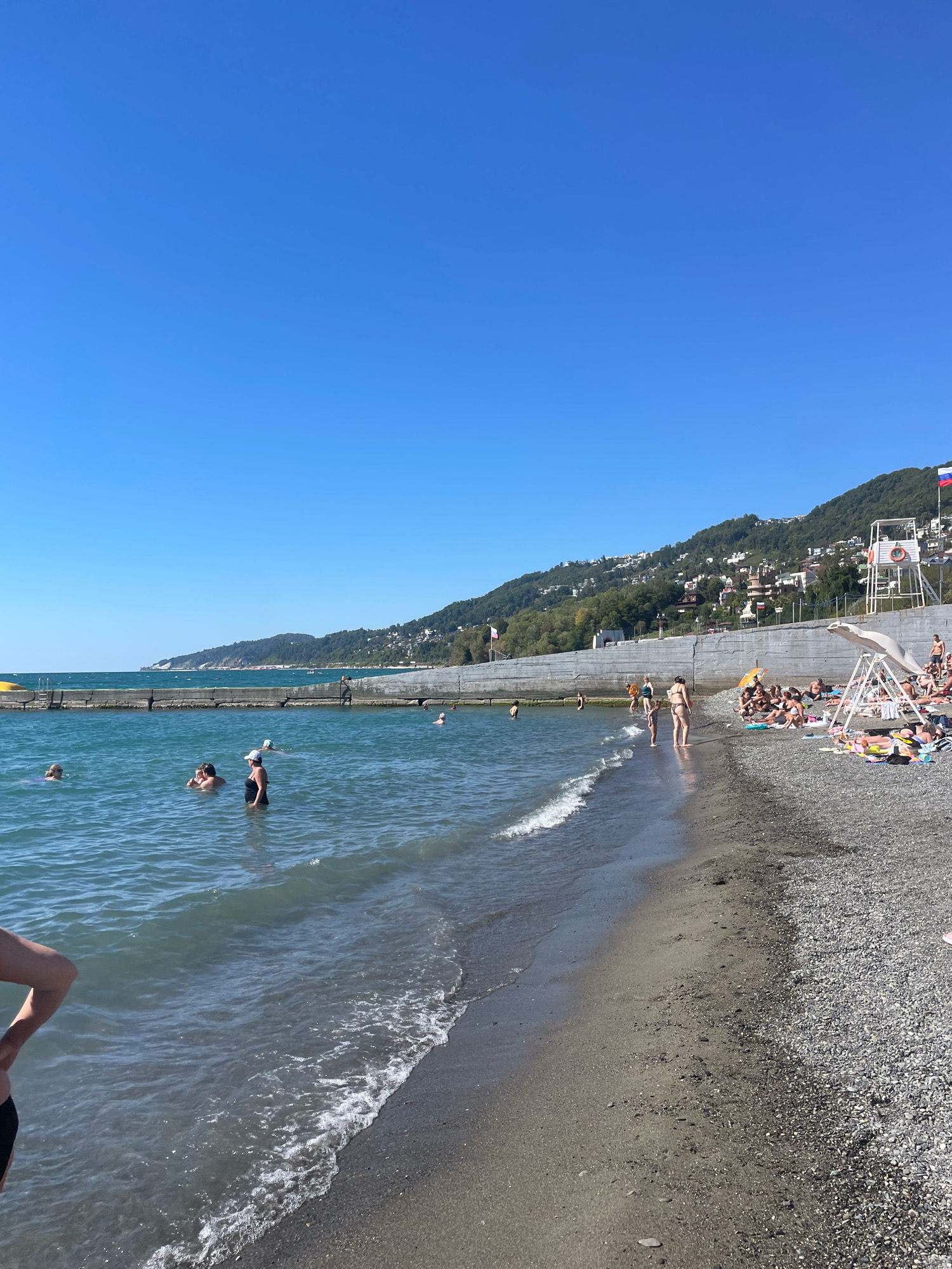 On a clear day, a pebble beach borders the Black Sea in Sochi, Russia. A long concrete embankment runs along the coastline, with a resort town nestled on the forested hillside above. Several people are in the water, including a group swimming near the embankment and individuals wading in the shallows. On the beach, a lifeguard tower stands tall, and numerous beachgoers are sunbathing on towels or relaxing under a beach umbrella. A Russian flag flies from the top of the lifeguard tower.