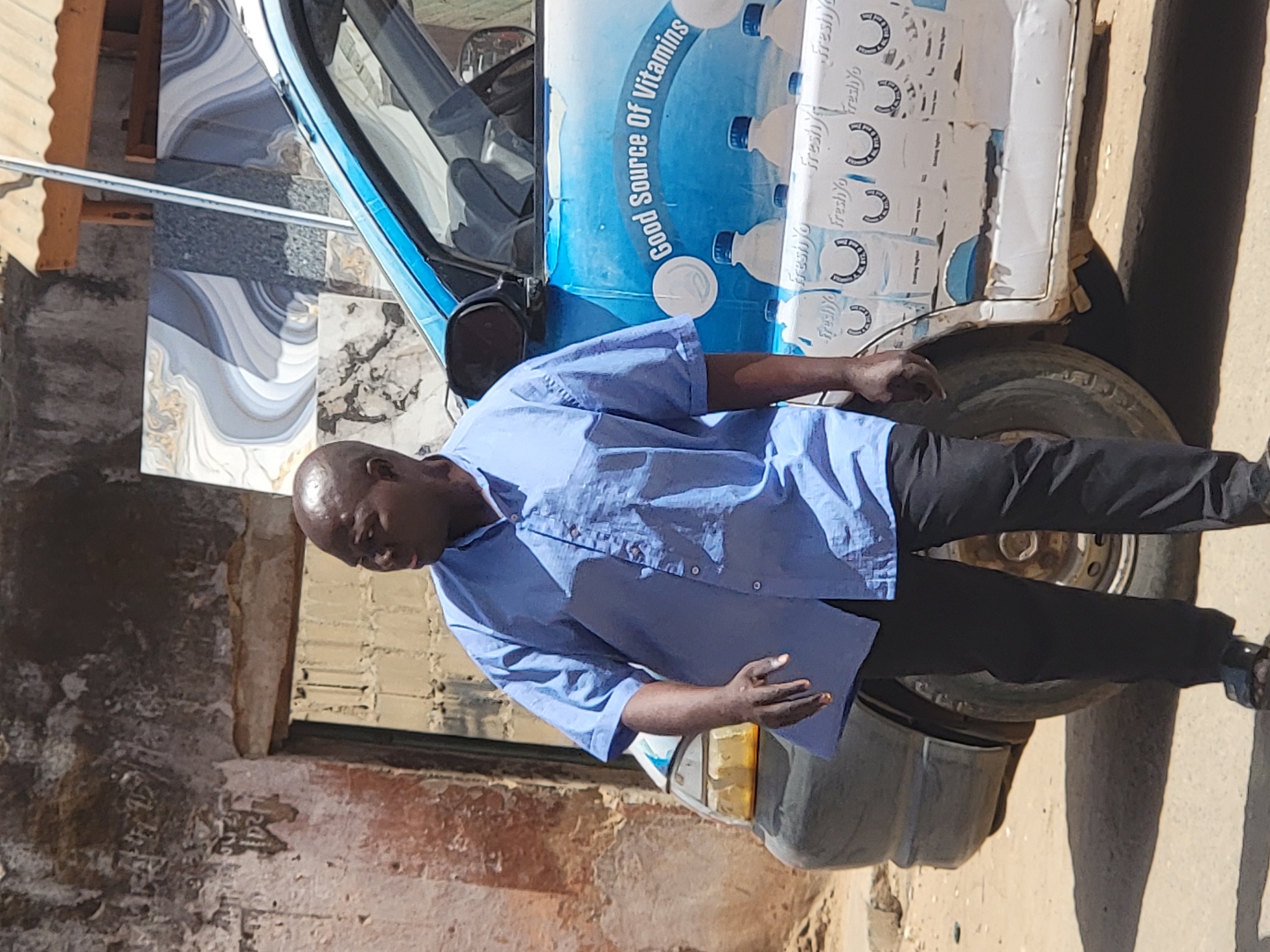A man wearing a short-sleeved blue shirt and black pants stands next to a parked vehicle. The vehicle's door is adorned with advertisements for "FreshYo" and the text "Good Source of Vitamins." The man appears to be holding a document or folder. The scene is situated outdoors, with an adjacent building or structure showing signs of age and wear. The ground is dirt or unpaved. The location is inferred to be in Maiburiji, Nigeria.
