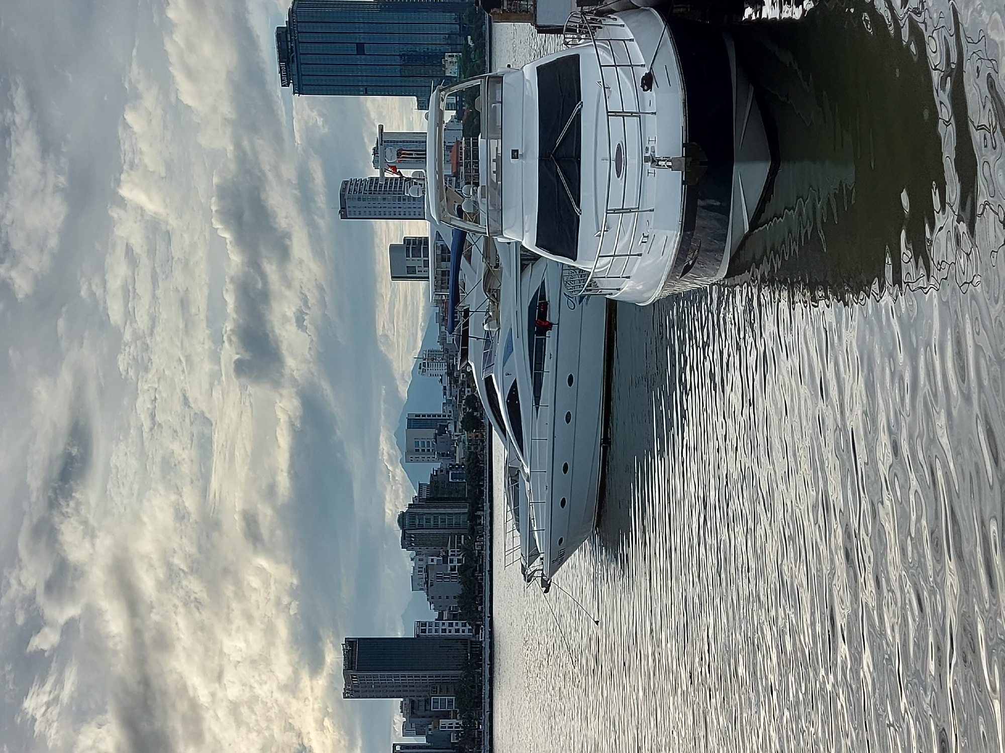 The image depicts a waterfront scene in the city of Đà Nẵng, Vietnam, likely along the Han River. In the foreground, two white yachts are moored, their sleek designs contrasting with the rippling, grey water. The yacht on the right is closer to the viewer and is docked at the edge of a pier, its dark hull visible beneath the waterline. The yacht on the left is slightly further back, its bow pointed towards the left of the frame.
The background features a cityscape with several modern high-rise buildings, including a prominent dark blue skyscraper with the "Marriott" logo visible near its top. Behind the buildings, the hazy outlines of mountains can be seen against a sky filled with dramatic, overcast clouds. The overall lighting suggests it is either early morning or late afternoon, with soft, diffused light. There are no visible people in the image.