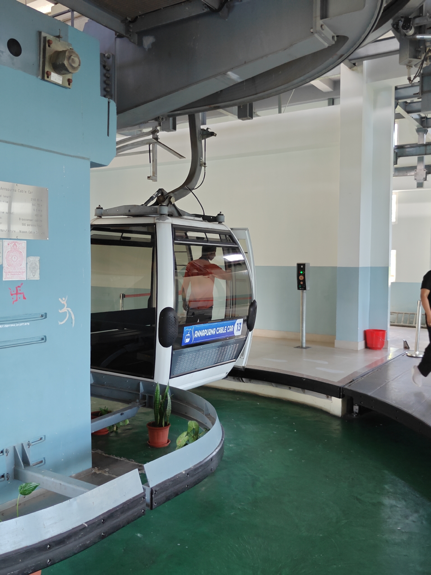The image depicts the interior of a cable car station in Pokhara, Nepal, specifically at the Annapurna Cable Car. A white and gray cable car, labeled "Annapurna Cable Car 13", is positioned on a track. Inside the cable car, a person wearing an orange shirt is visible. To the left of the cable car, a light blue pillar displays technical specifications and two religious symbols. Potted plants are placed along the curved track. In the background, a traffic light signals operation, and a red bucket sits near a wall. The floor is a dark green surface. The overall environment appears to be a functional station with elements of both industrial infrastructure and decorative greenery.