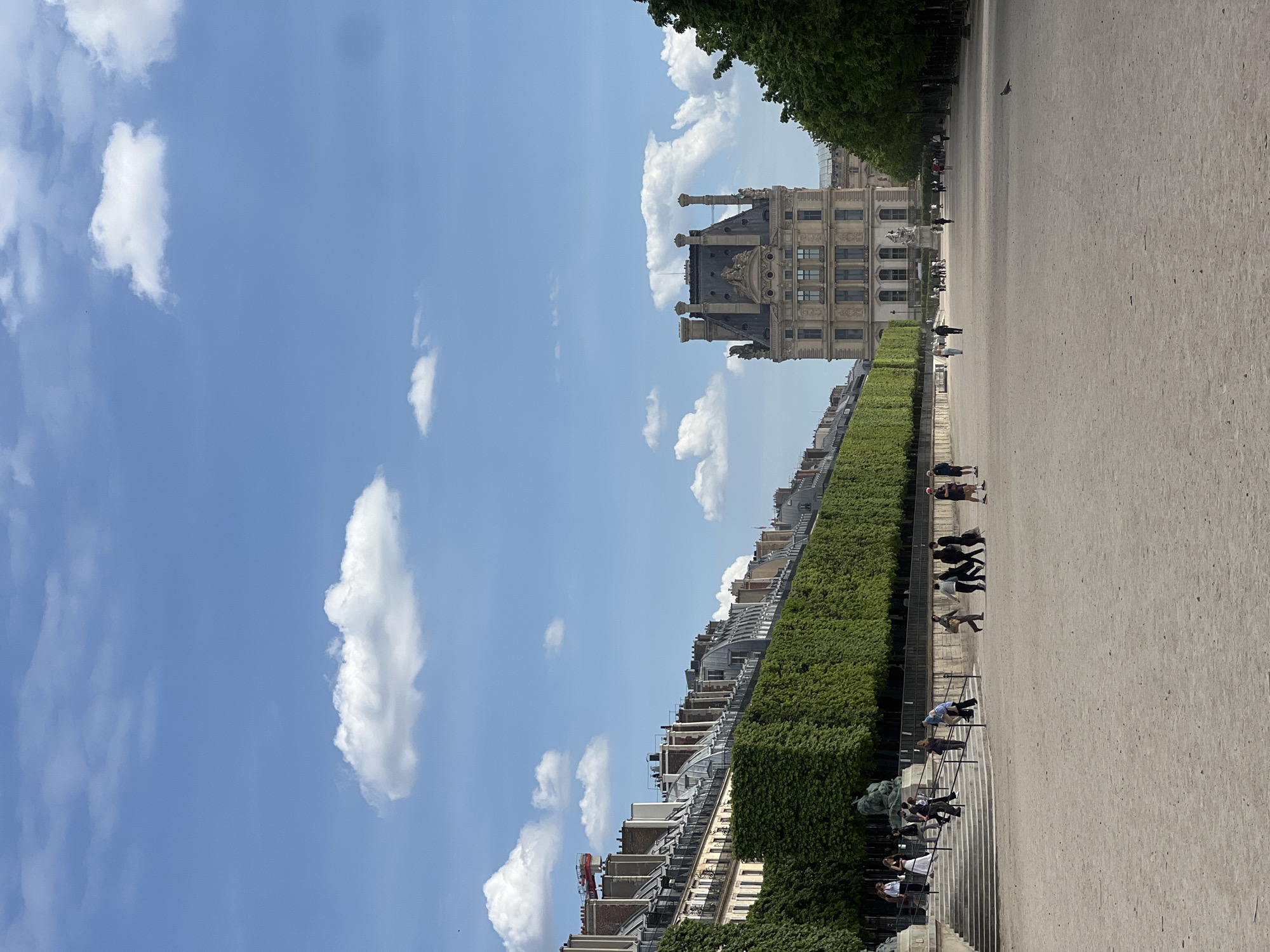 The image displays a panoramic outdoor scene under a clear, bright blue sky with scattered white cumulus clouds. The perspective is wide, capturing a large, open space characteristic of a Parisian garden or public square.
In the mid-ground and extending towards the background, a grand, ornate building stands prominently, exhibiting classical architecture with multiple windows, decorative sculptures, and distinctive roof structures including turrets and chimneys. This building is a strong architectural cue for the Louvre Museum.
Across the foreground and stretching into the distance is a broad, light-colored path, likely composed of gravel or dirt. This path is bordered by a long, neatly trimmed green hedge. Beyond the hedge, a row of urban buildings with numerous windows and balconies extends along the horizon. On the far side of the path, a line of tall, leafy green trees forms a natural boundary.
Several people are scattered throughout the scene. Some are casually walking or standing on the path, while others are seated on steps or benches near the green hedge. Their activities suggest a leisurely enjoyment of the outdoor environment. A single bird, possibly a pigeon, is visible on the distant path.
The bright sunlight and relatively short shadows indicate it is a clear, sunny day, likely late morning or early afternoon. The overall atmosphere appears peaceful and relaxed. There is no visible text in the image. The architectural style and formal landscaping strongly suggest the location is in Paris, France, consistent with the context provided.