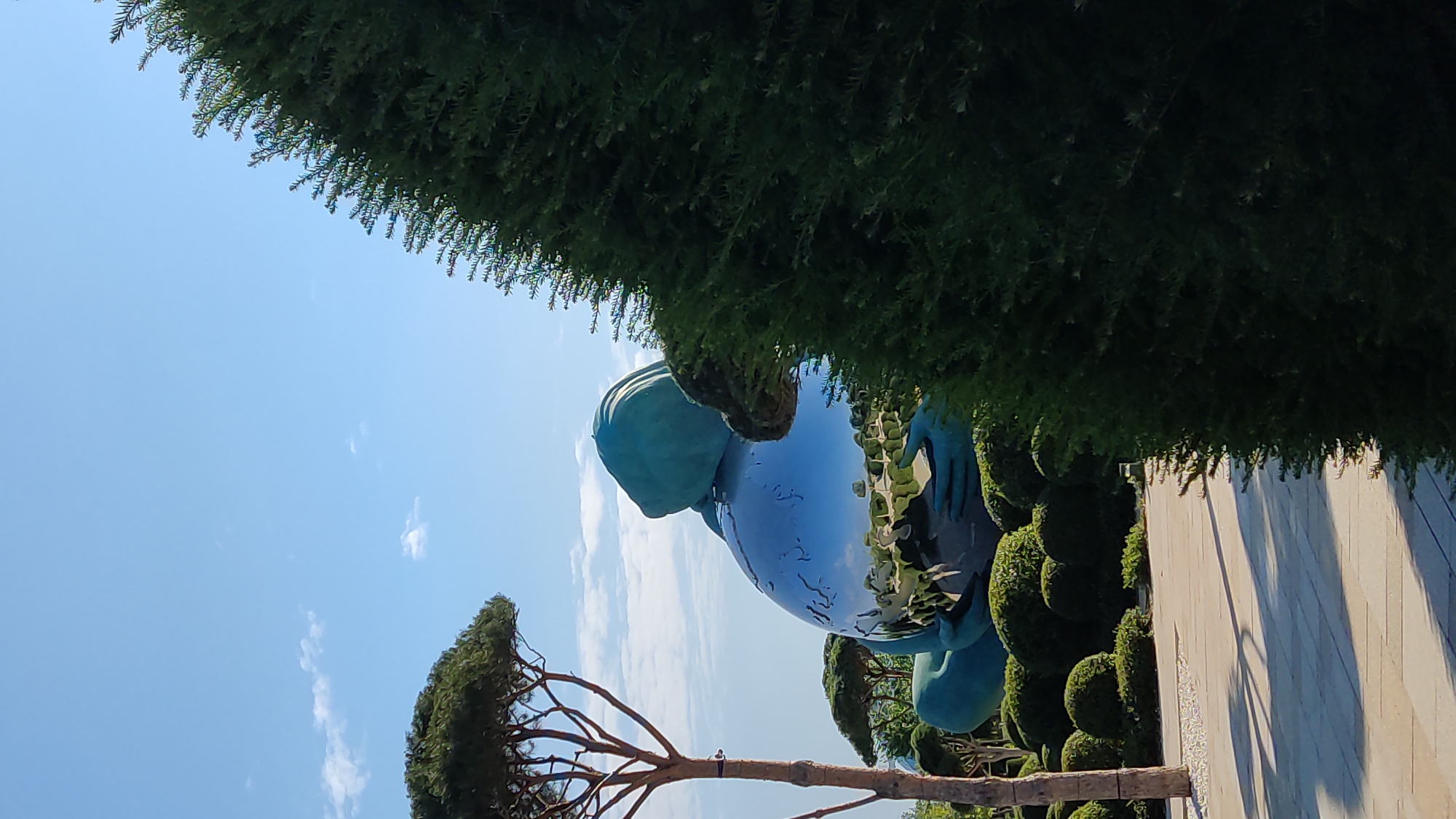 The image depicts a large, teal-colored sculpture of a figure holding a reflective sphere, resembling the Earth. The sculpture is situated outdoors in a landscaped garden area. The sphere reflects the surrounding environment, including a blue sky with scattered clouds, trees, and manicured bushes. A tall, slender tree with a dark trunk stands to the left of the sculpture. In the foreground, a paved pathway with dappled sunlight and shadows runs horizontally across the image. A dense, dark green evergreen bush is prominently positioned on the right side, partially obscuring the sculpture. This sculpture is located in Krasnodar, Russia, specifically in the "Galitsky Park," a well-known public park.