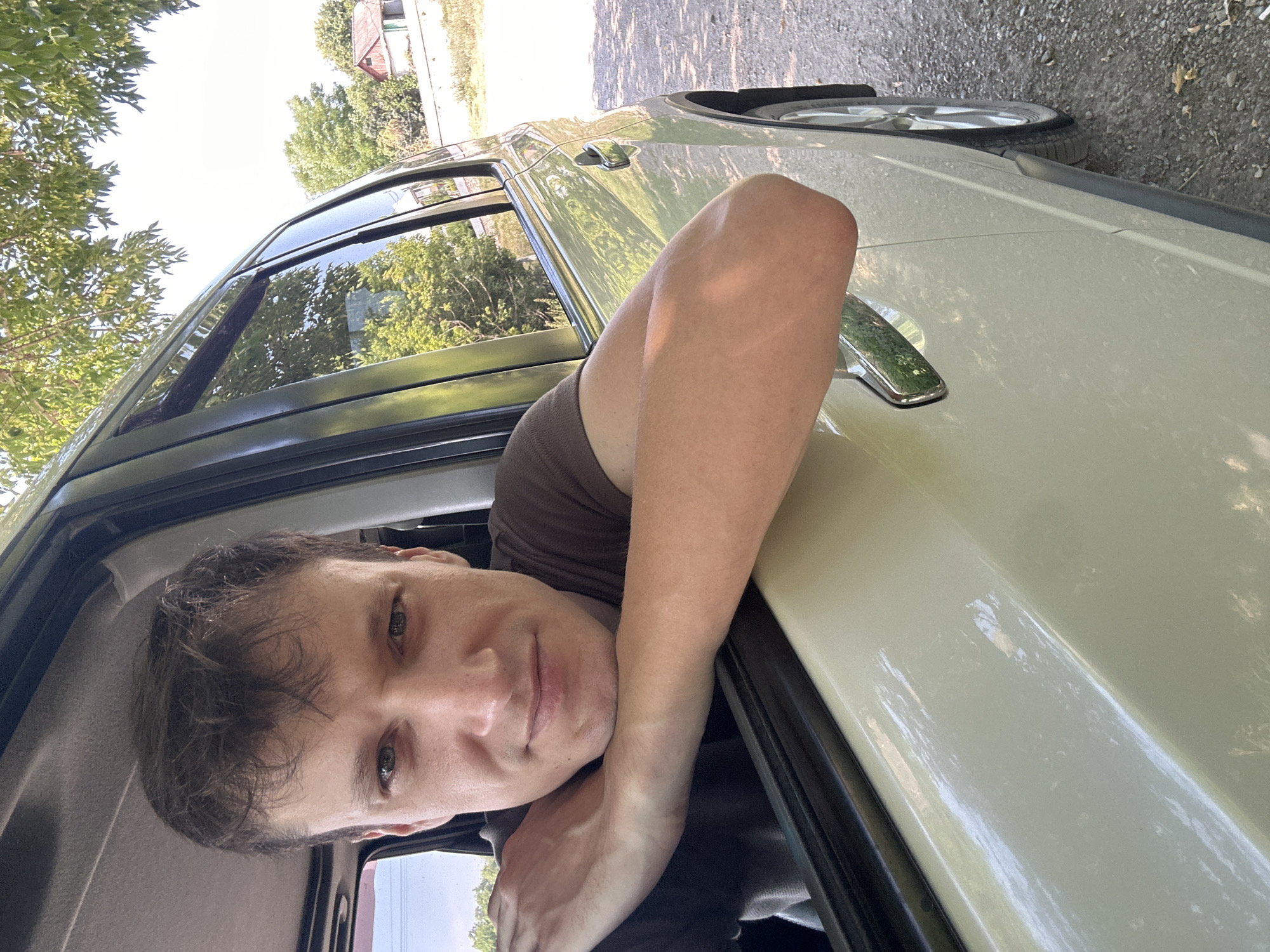 A man is leaning out of a car window, resting his arms on the windowsill. The car is light-colored and appears to be parked on a gravel road, possibly in a rural area outside of Makiv, Ukraine, given the surrounding trees and a small building visible in the background. The man has light skin and short, dark hair. He is wearing a brown t-shirt. The sun is shining, casting shadows and highlighting the car's glossy surface. The scene suggests a casual moment captured outdoors, likely during the daytime.