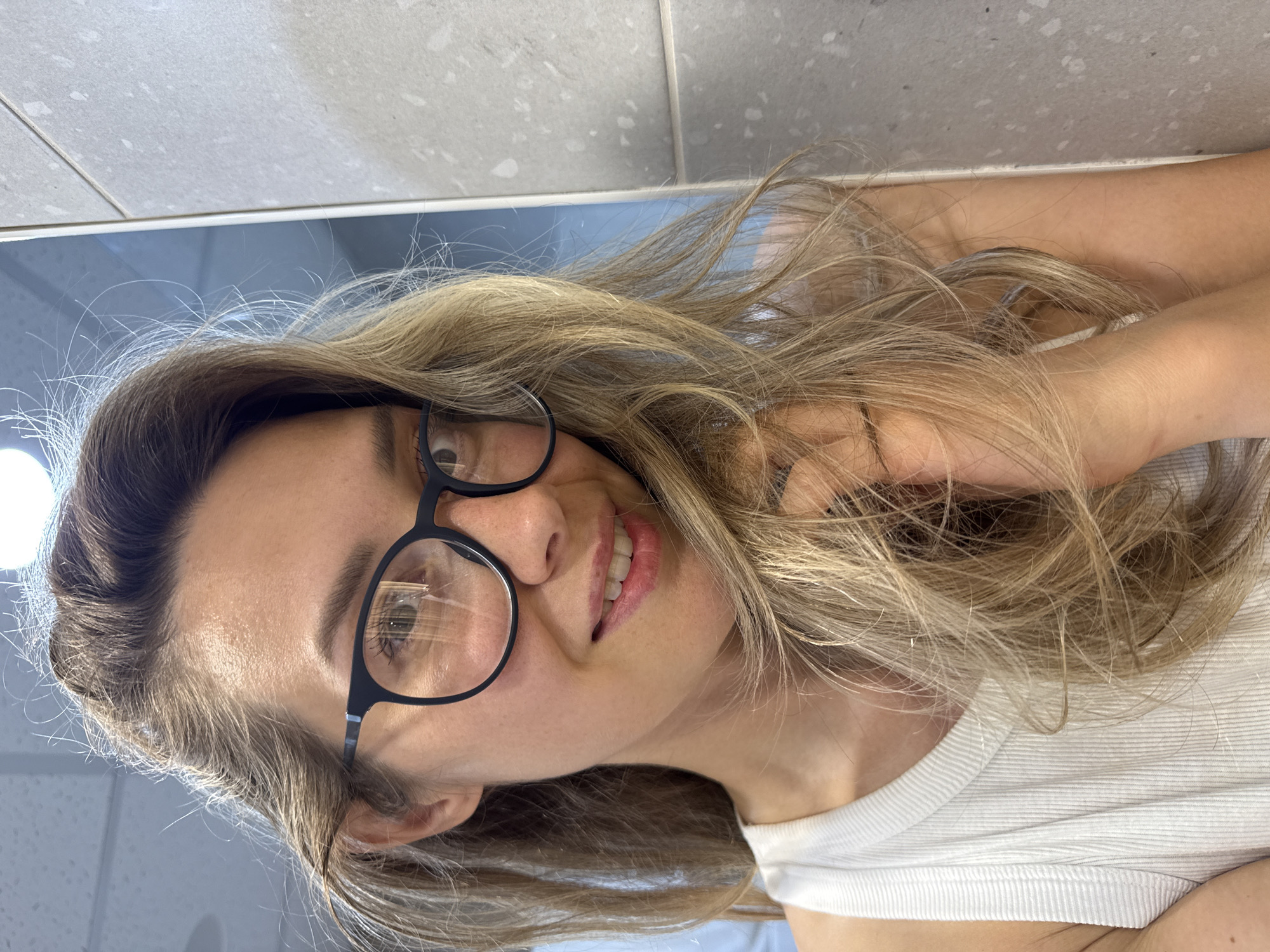 The image shows a close-up, selfie-style shot of a young woman with long, blonde, wavy hair. She is wearing black-rimmed glasses and a white, ribbed tank top. Her hair is falling over her face and shoulder. She is smiling gently. The background is blurred, but appears to be an indoor setting with a tiled wall to the right and a light-colored ceiling with textured tiles above. No specific location in Nebug, Russia, can be identified from the image.