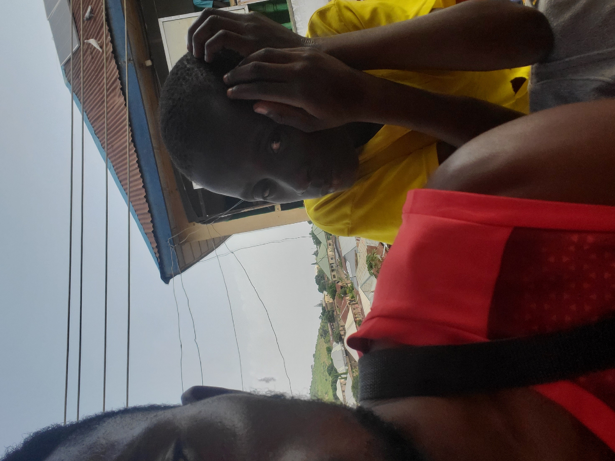 The image shows a close-up of two individuals. The person on the left is partially visible from the shoulders up, wearing a red garment. Their left side of their face and hair are visible. The person on the right is a young individual wearing a yellow t-shirt, with their head resting on their hands. They are looking to the left. The background shows a corrugated metal roof with solar panels and two pigeons. Beyond that, a hilly landscape with buildings, indicative of a town or city, is visible. This scene is consistent with a location in the city of Gwong, Nigeria, characterized by its typical urban and semi-urban architecture, and the presence of natural hills in the surroundings. The sky is overcast.