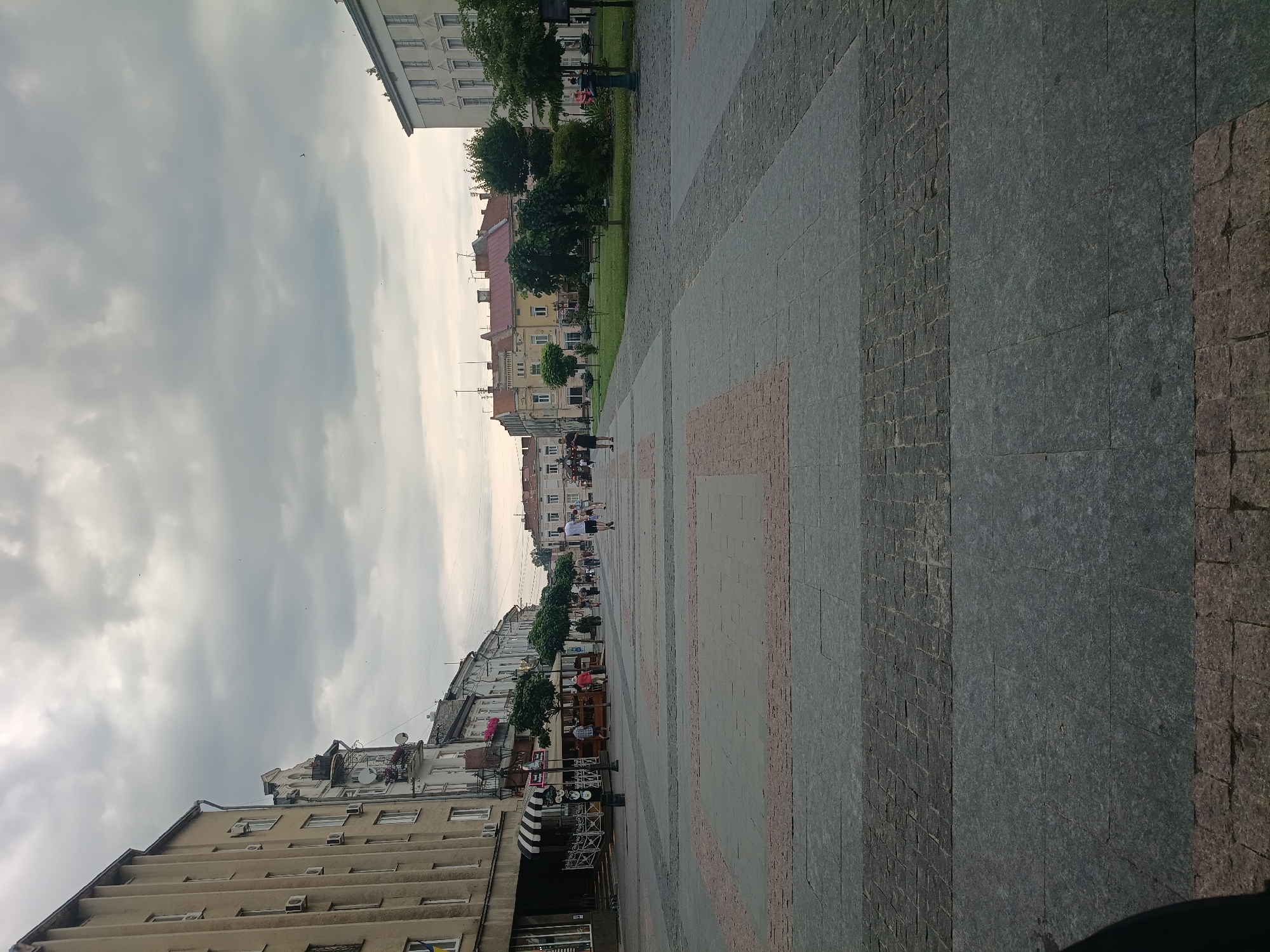 The image shows a view of a paved pedestrian area in front of buildings in Drohobych, Ukraine. The paving is made of large, grey flagstones with rectangular sections of pinkish-orange and smaller grey cobblestones interspersed. The area is lined with trees and a grassy area with benches. Several people are walking in the distance on the pedestrian area and some are seated in an outdoor cafe. The buildings on the left are multi-story with a light yellow facade and windows. The buildings across the street are of various architectural styles and colors, with pitched roofs. The sky is overcast with grey clouds.