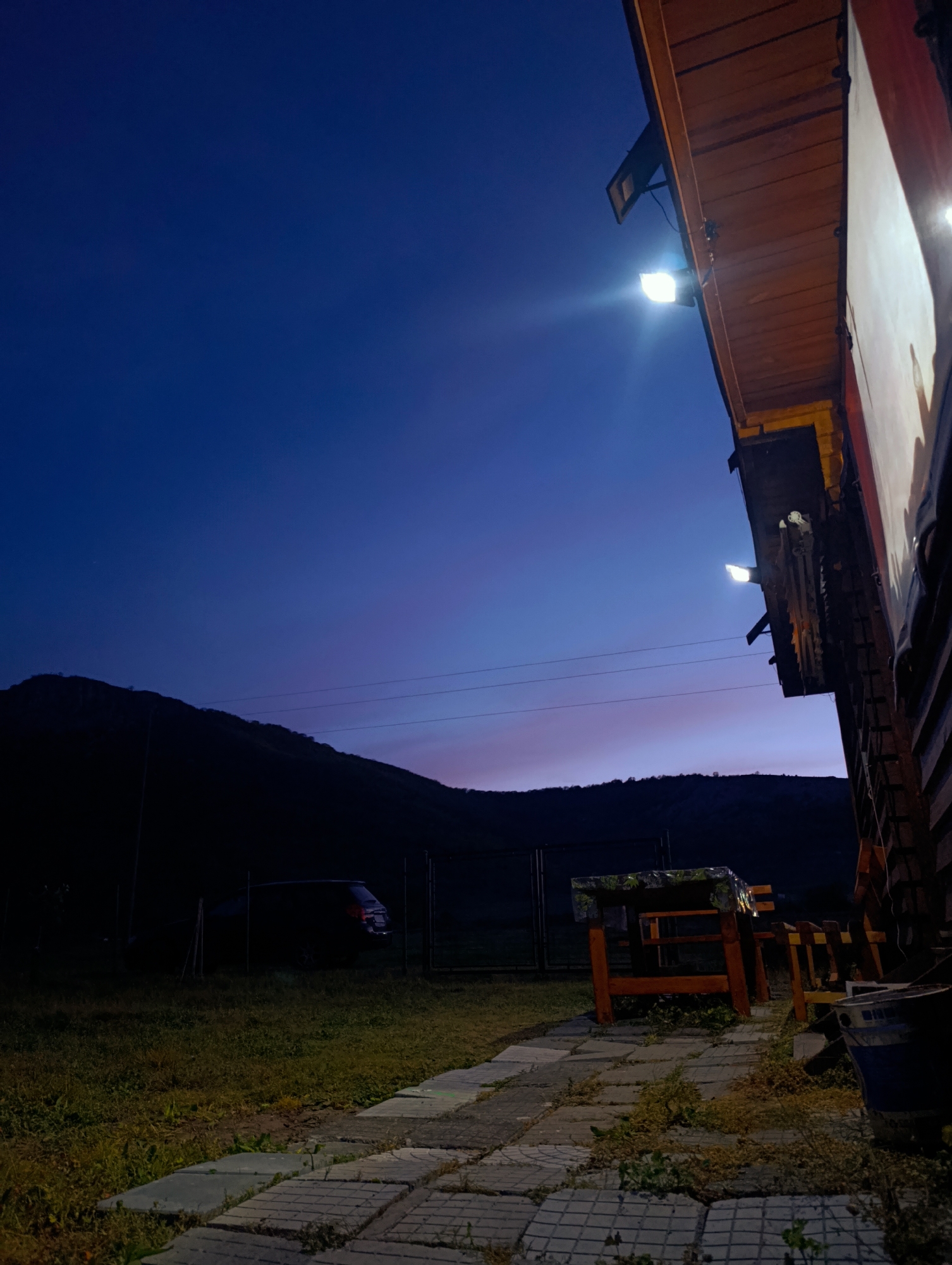 The image depicts an outdoor scene at dusk, possibly near or in the city of Shiroko pole, Bulgaria, given the mountain in the background and the architecture. A dark mountain silhouette is visible in the distance beneath a dark blue and pink sky. A car is parked in the grass. In the foreground, a brick path leads to a wooden table and benches next to a wooden building. Two lights affixed to the building illuminate the area. There are miscellaneous objects and a bucket visible in the lower right corner near the building's edge.