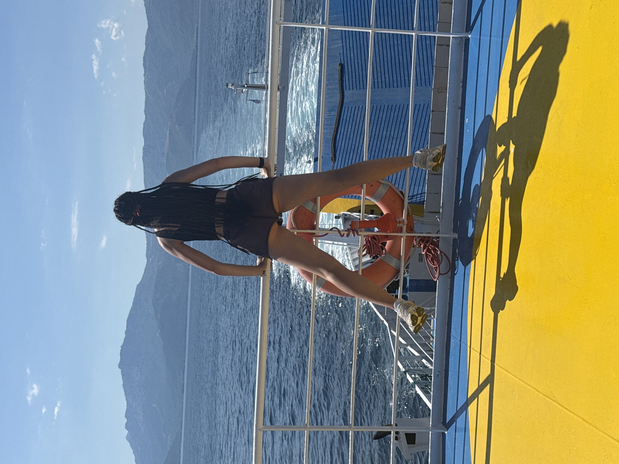 A person with long, dark braids stands on the deck of a moving ferry, facing away from the camera and looking out at a body of water. The person is wearing a black crop top and dark shorts, with white sneakers. They are leaning on a white railing, with their legs spread apart and slightly bent. An orange life preserver is attached to the railing behind them. The ferry deck is painted yellow and blue. In the background, mountains are visible across the water under a clear blue sky. The ferry is creating a wake in the water behind it. The scene appears to be in Greece, though the specific city is unknown.