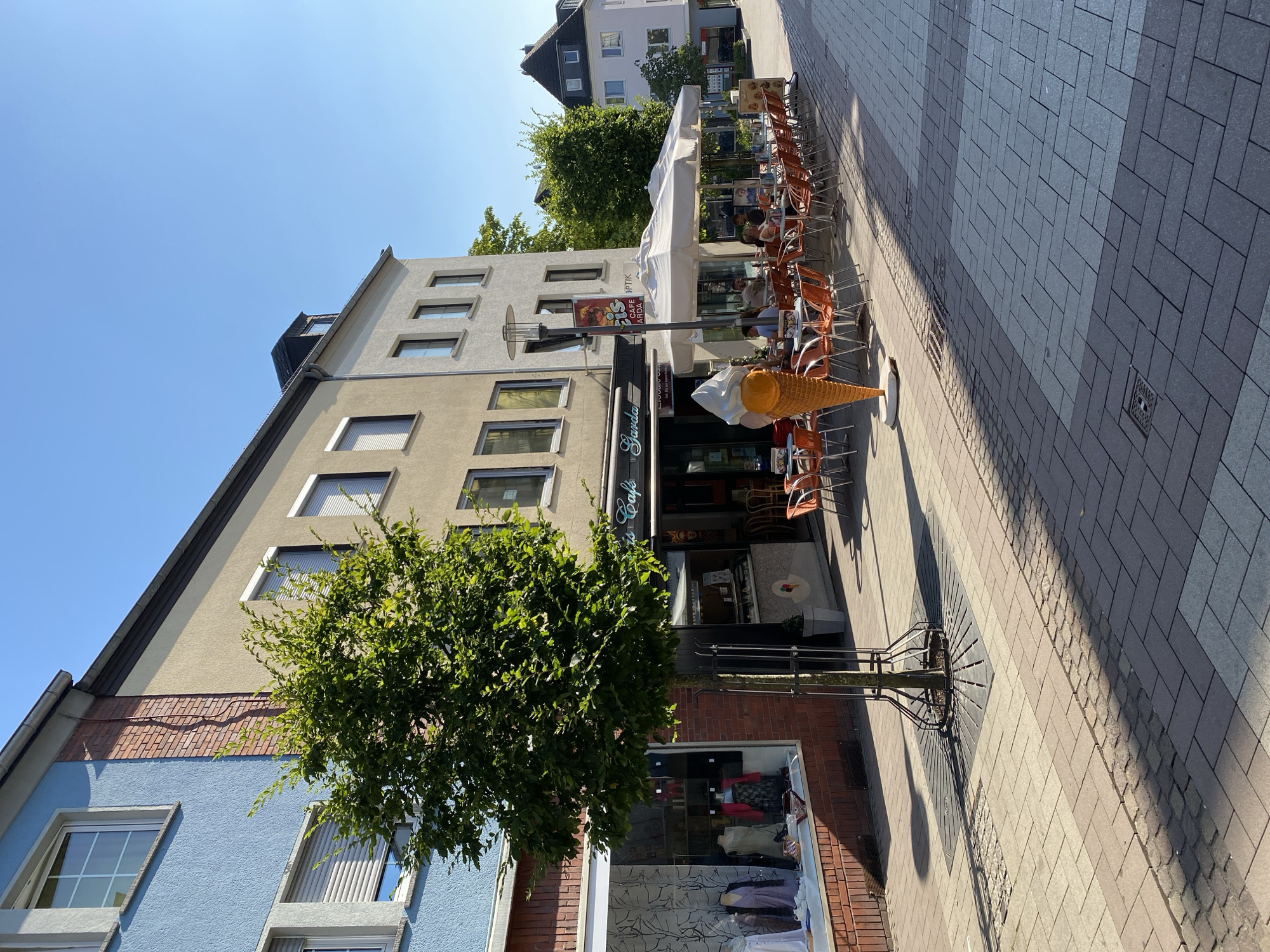 The image depicts an outdoor scene in Altenkirchen, Germany, on a sunny day. The perspective is angled, looking down a street lined with buildings and a sidewalk. In the foreground, a large, decorative ice cream cone statue stands on the paved sidewalk. To the left, a multi-story building features a blue facade on its lower levels and a lighter beige facade on its upper levels, with several windows visible. A lush green tree with dense foliage is positioned in front of this building, casting a shadow on the sidewalk.
To the right of the tree, the sidewalk extends into the distance, paved with rectangular stones. A row of outdoor seating for a cafe is set up along the side of a building, with orange chairs and small tables. An open white umbrella shades some of the tables. Several people are seated at the tables, appearing to be patrons of the cafe. The building behind the cafe has large windows and signage. One sign, partially obscured, includes the word "Cafe" and some German text. Another sign above an entrance reads "Garda". Further down the street, more buildings and cafes are visible, with people walking and sitting. The time of day appears to be midday, given the bright sunlight and distinct shadows. The overall atmosphere is relaxed and suggests a pleasant day for outdoor dining and strolling in a town setting.