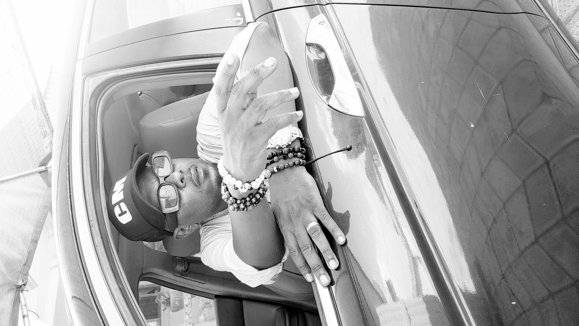 A black and white image shows a man partially inside the trunk of a dark car, with the car's door open. He is wearing a backwards baseball cap with "GM" visible, square sunglasses, a white t-shirt, and several bracelets on his left wrist. His right hand rests on the car door. The interior of the trunk is visible, with a light-colored seat. The background is blurry, suggesting an outdoor setting possibly in the city of Sabon Tasha, Nigeria.
