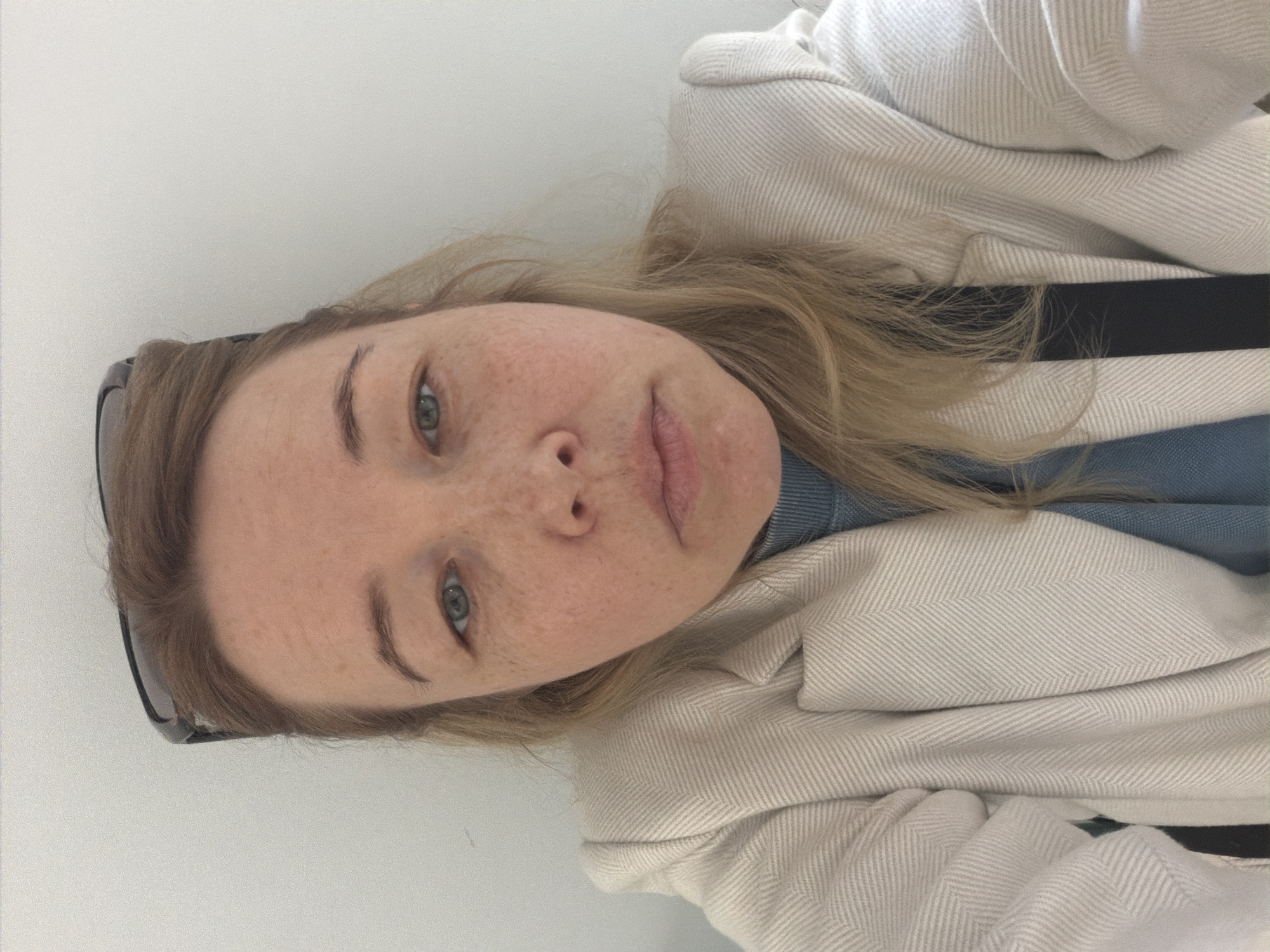 The image shows a woman's face, angled slightly. She has fair skin, freckles, light blonde hair, and gray-blue eyes. Sunglasses are perched on top of her head. She is wearing a light-colored coat over a blue shirt or sweater, and a black strap is visible. The background is a plain, light-colored wall. No information indicates that the picture was taken in the city of Pervomaysky, Russia.